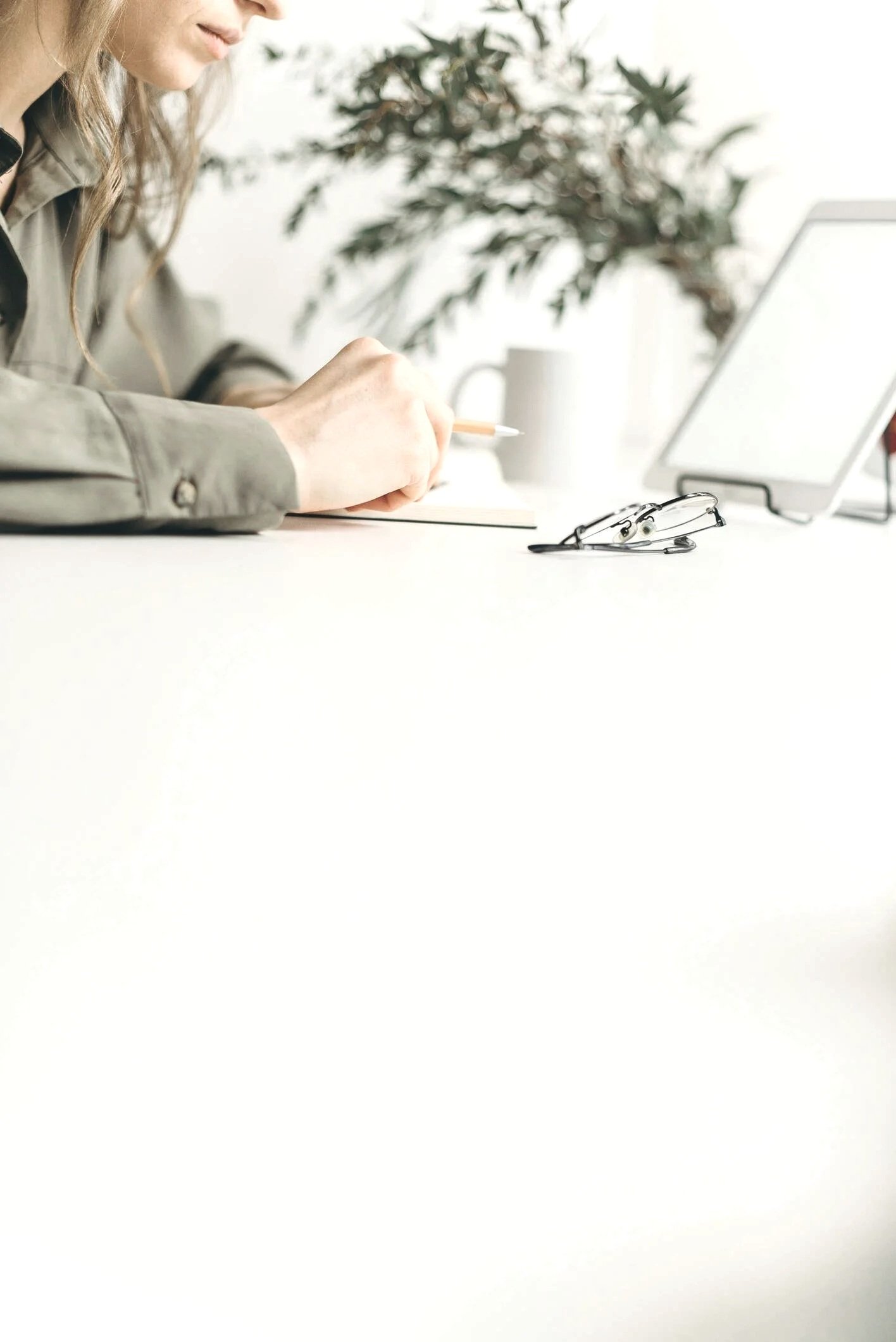 A woman sitting at a bright white desk, writing with a pen. On the desk, there are glasses, a white mug, and a computer monitor. In the background, a green plant is visible.