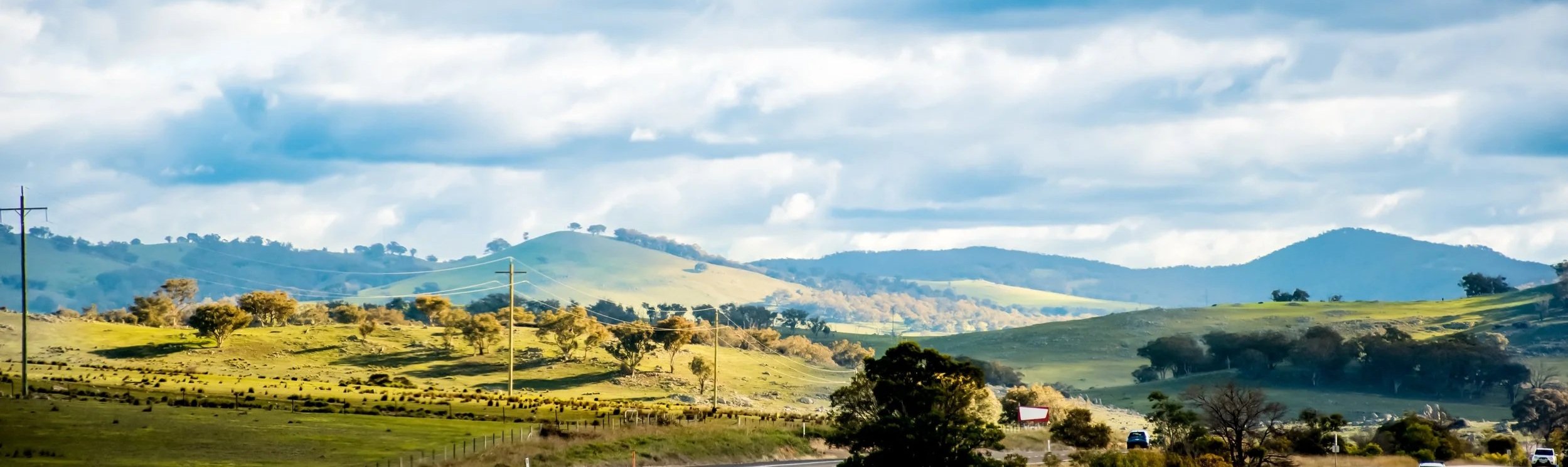 Rolling green hills stretch across the countryside beneath a broad sky, with scattered trees and distant ridgelines creating a layered rural landscape that conveys space, tranquillity and regional character.