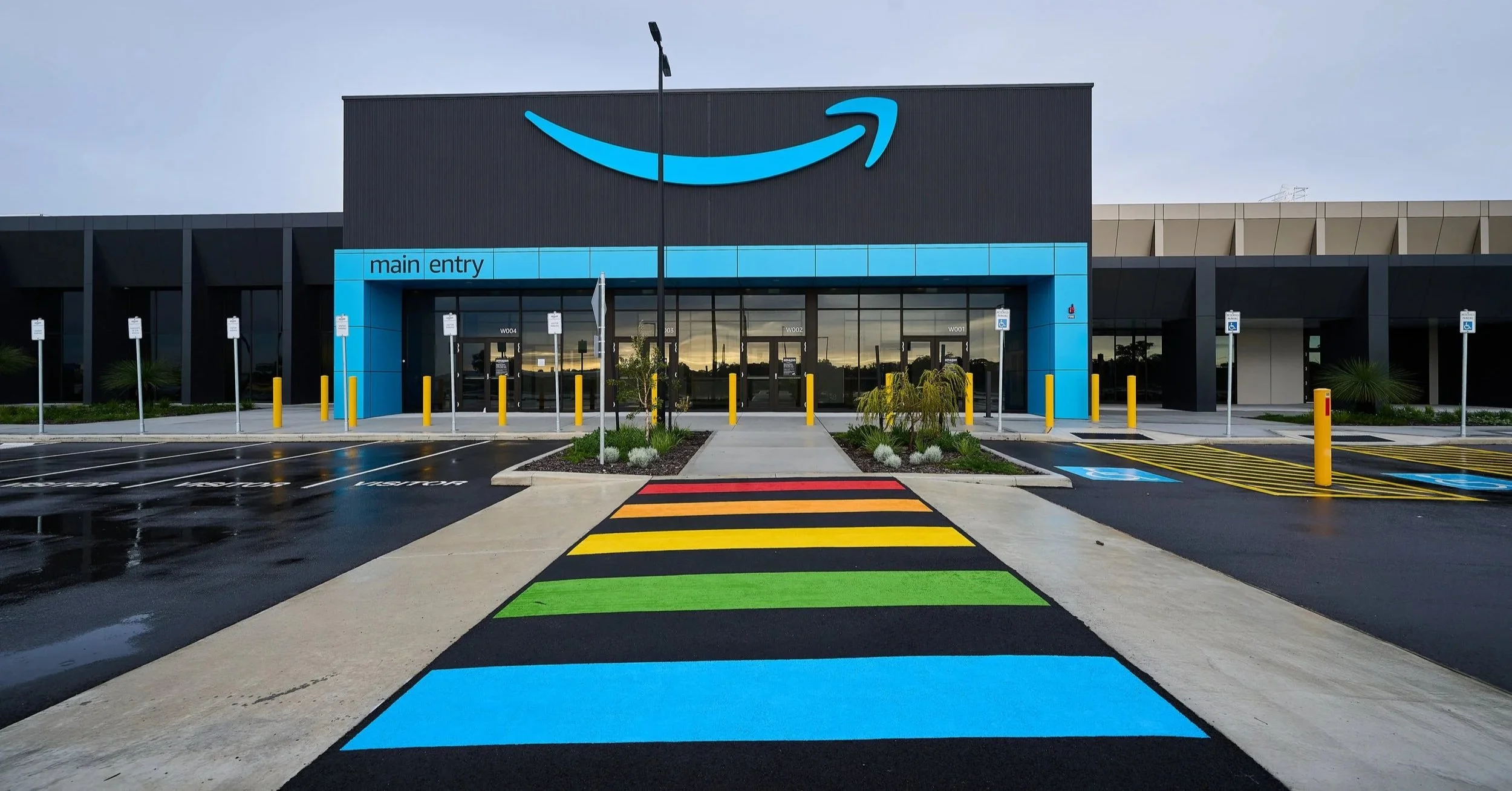 Main entrance to Amazon Australia’s PER4 fulfilment centre, with a rainbow pedestrian crossing, blue Amazon logo and accessible parking.