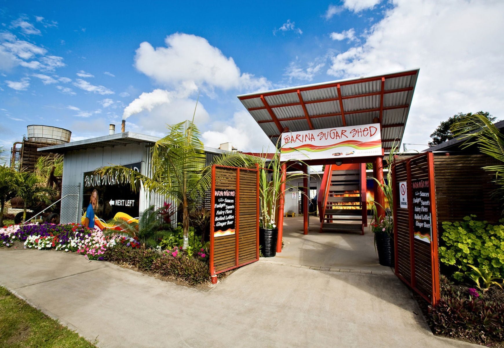Entrance of Sarina Sugar Shed with palms, flowers and sugar cane around the front gate.