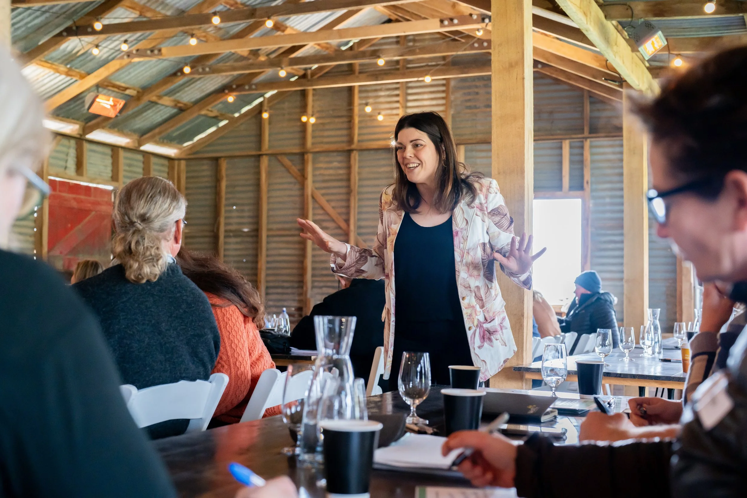 Giovanna Lever facilitates an engaging discussion with participants gathered around tables in a rustic workshop setting.