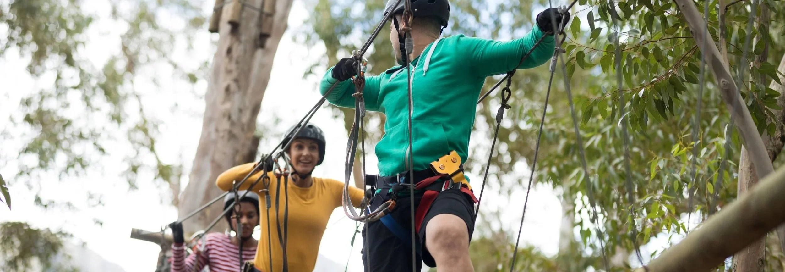 Two participants wearing helmets and safety harnesses navigate a high ropes course among trees, concentrating as they clip onto cables and balance along the elevated challenge.