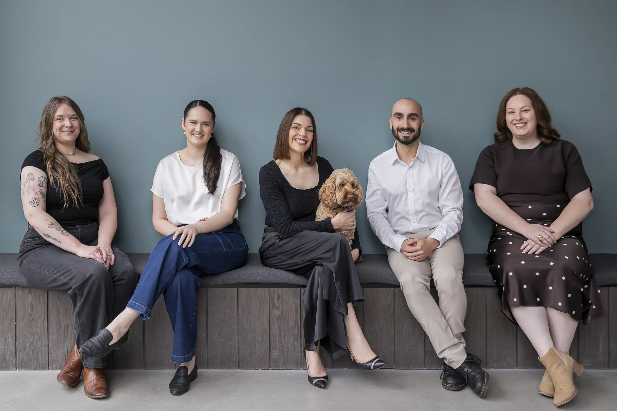 The Sparrowly Group team sits together smiling against a soft blue backdrop, joined by their office dog Anakin.