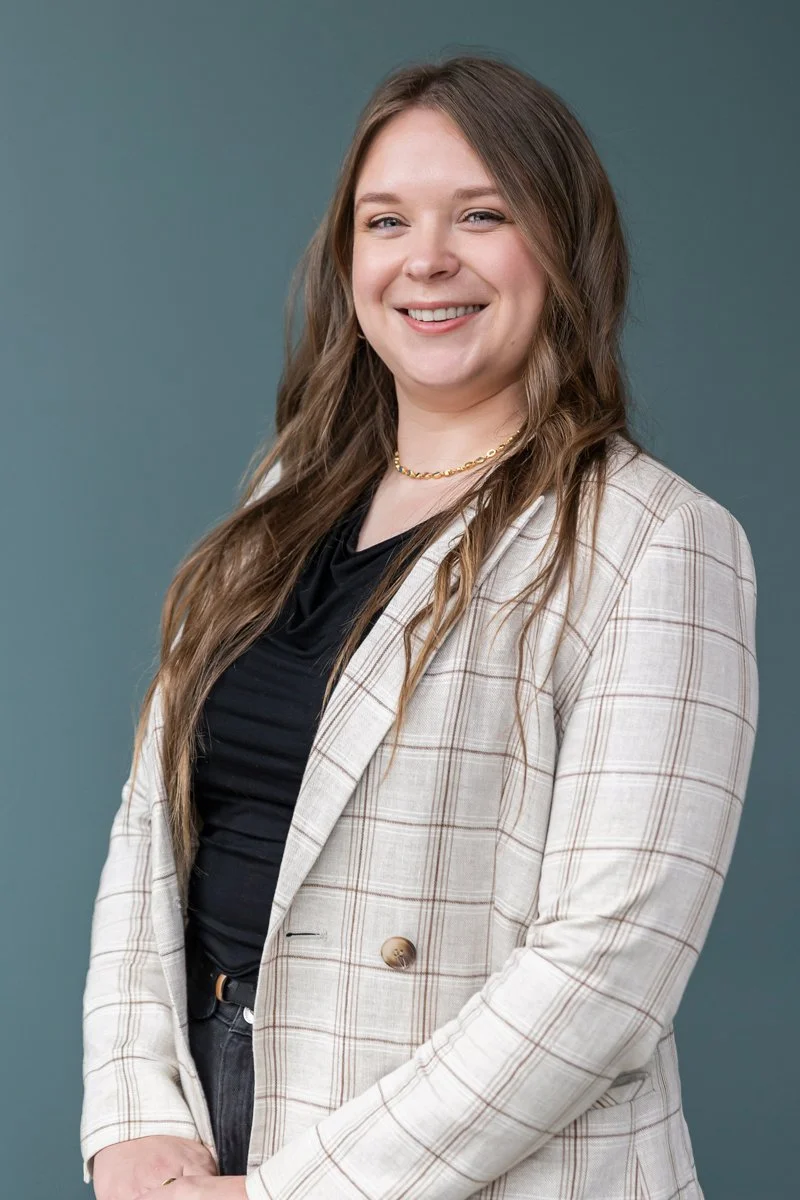 Portrait of Beth Burns smiling warmly, wearing a checked blazer over a black top against a soft blue background.