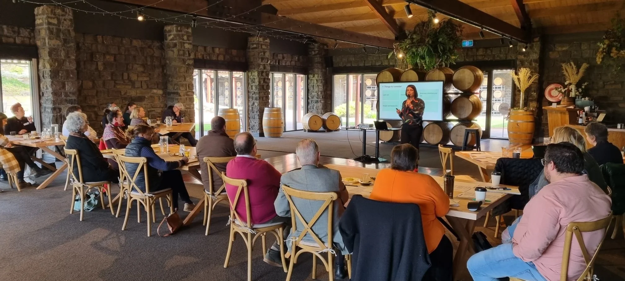 Giovanna Lever delivers an agritourism workshop at a winery, presenting to participants seated around communal tables in a barrel-lined venue, combining practical insights with an engaging, hands-on learning environment.