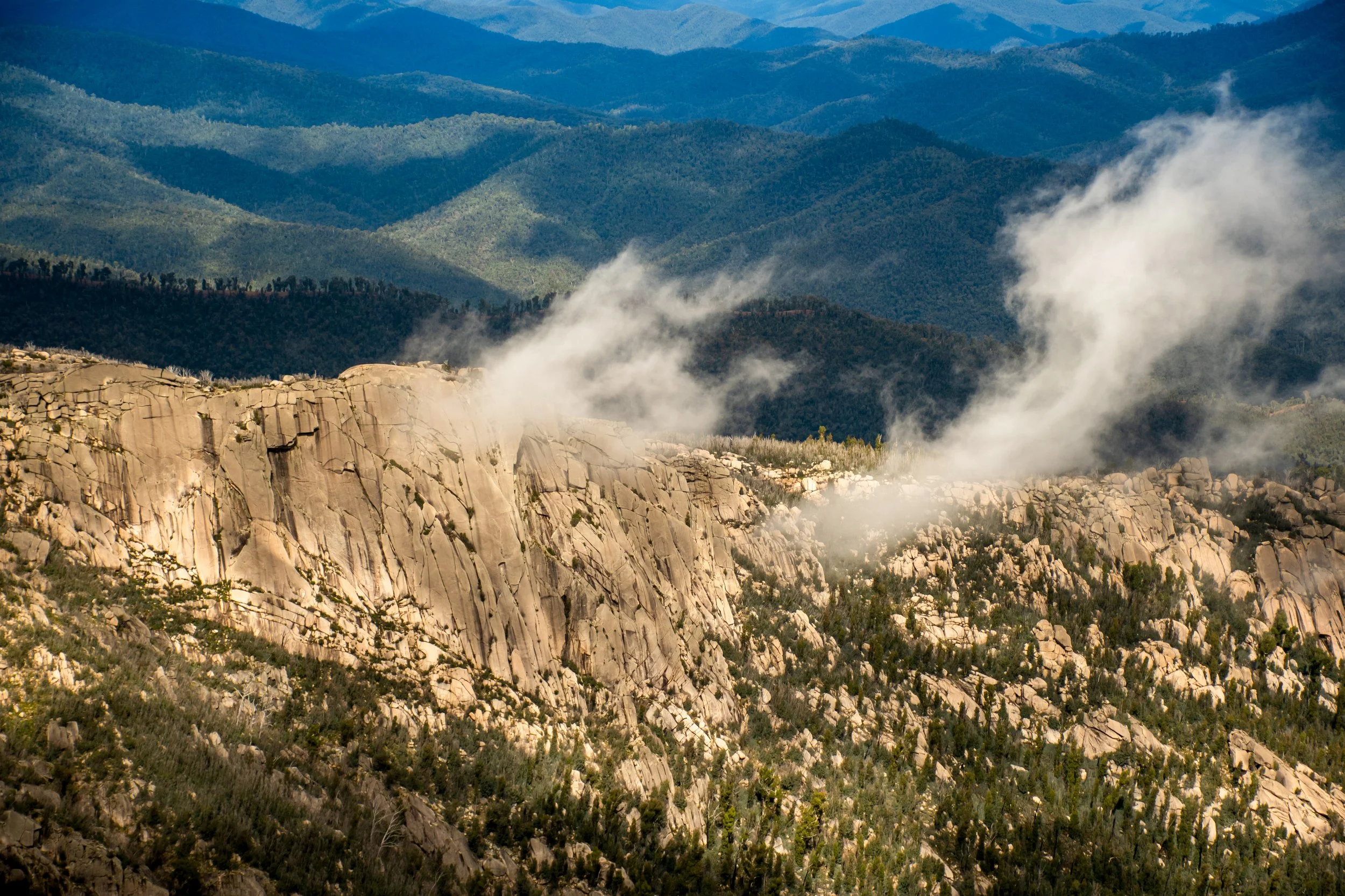 Steep granite cliffs rise from Mount Buffalo National Park in Victoria, their weathered rock faces partially veiled by drifting mist, with alpine vegetation clinging to the rugged plateau above and forested valleys stretching into the distance.