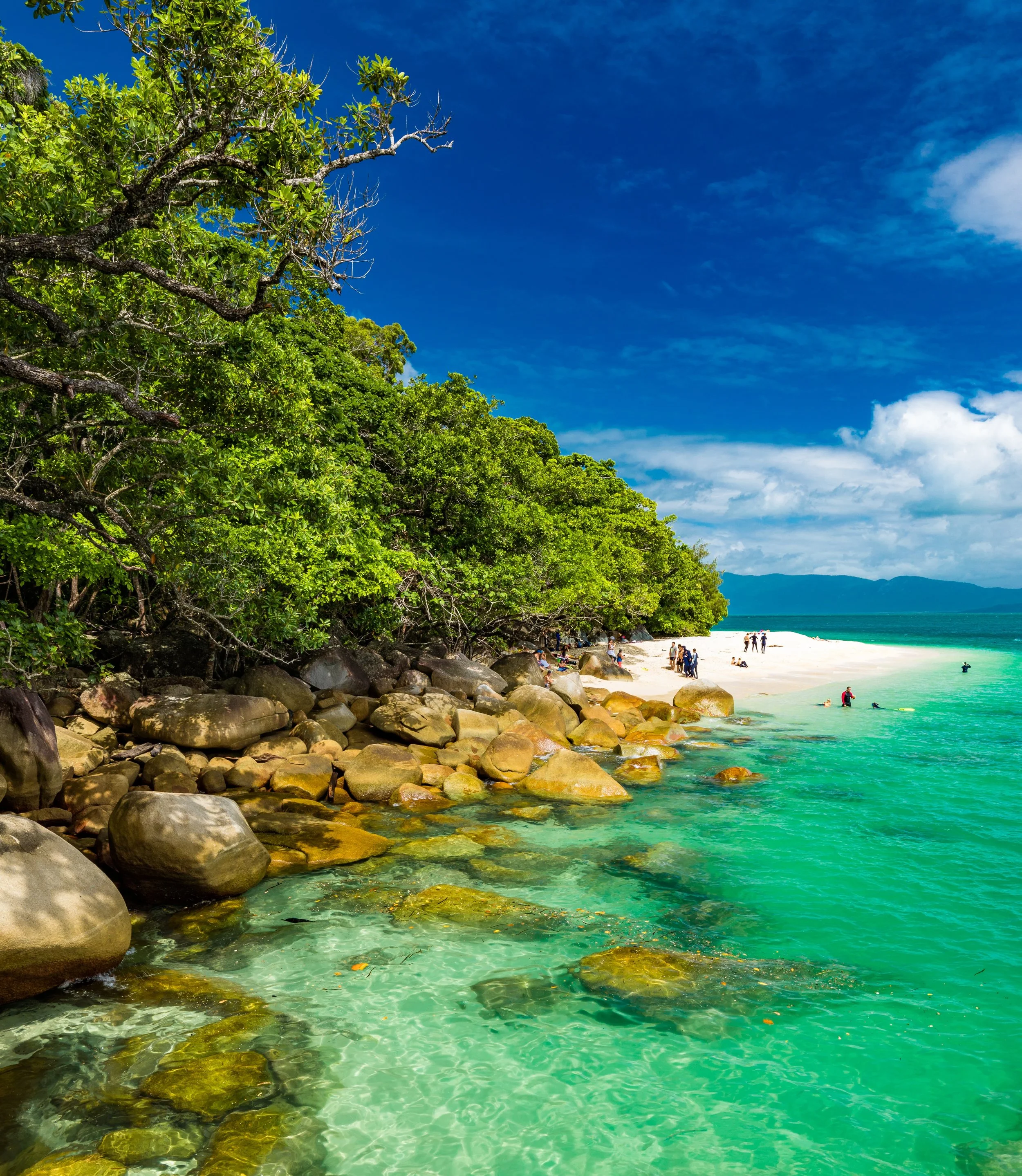 A clear teal beach with rocks, white sand, blue skies and shaded by green foliage.