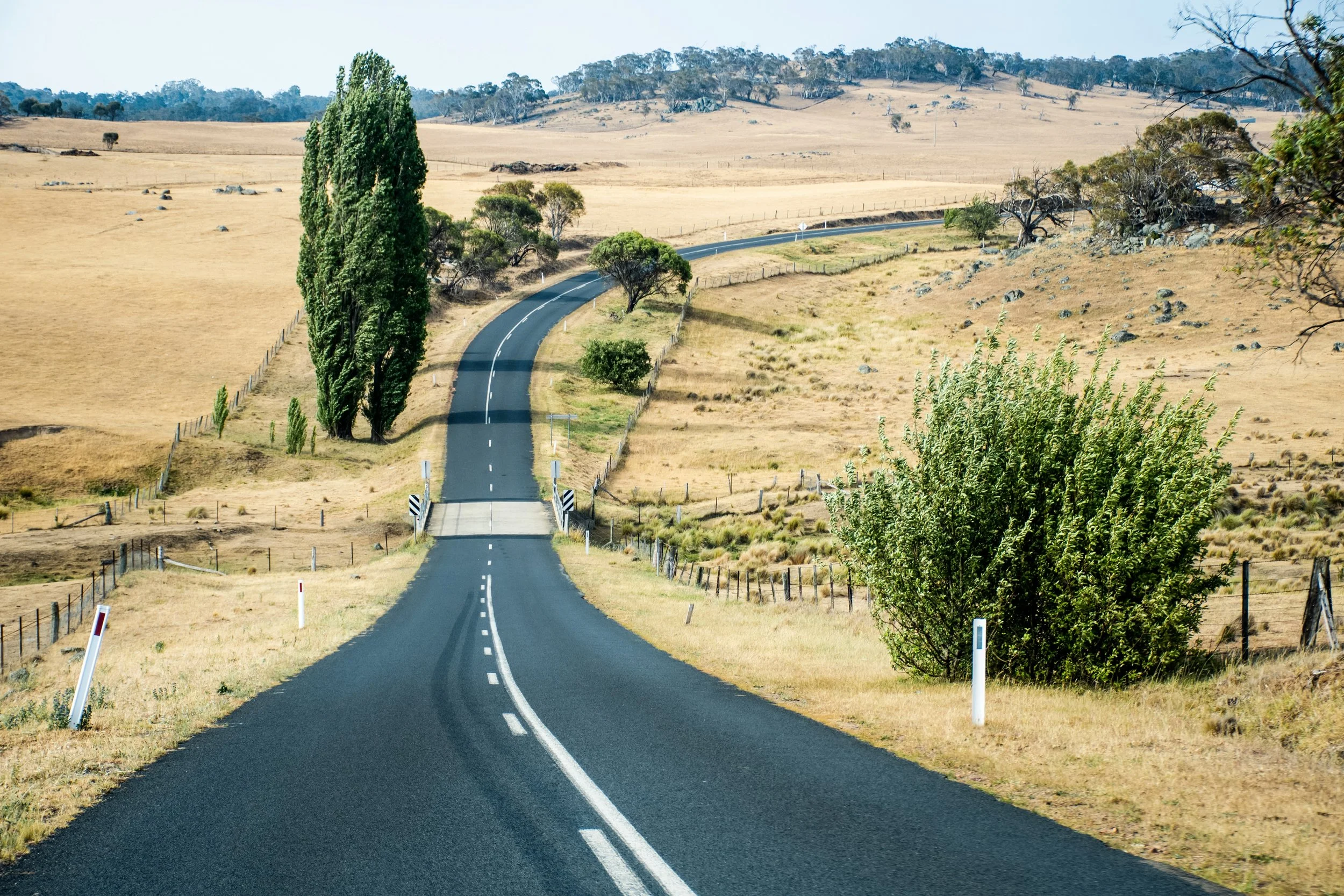 A winding country road stretches through dry rural farmland, symbolising regional connectivity, travel and the journey between communities.
