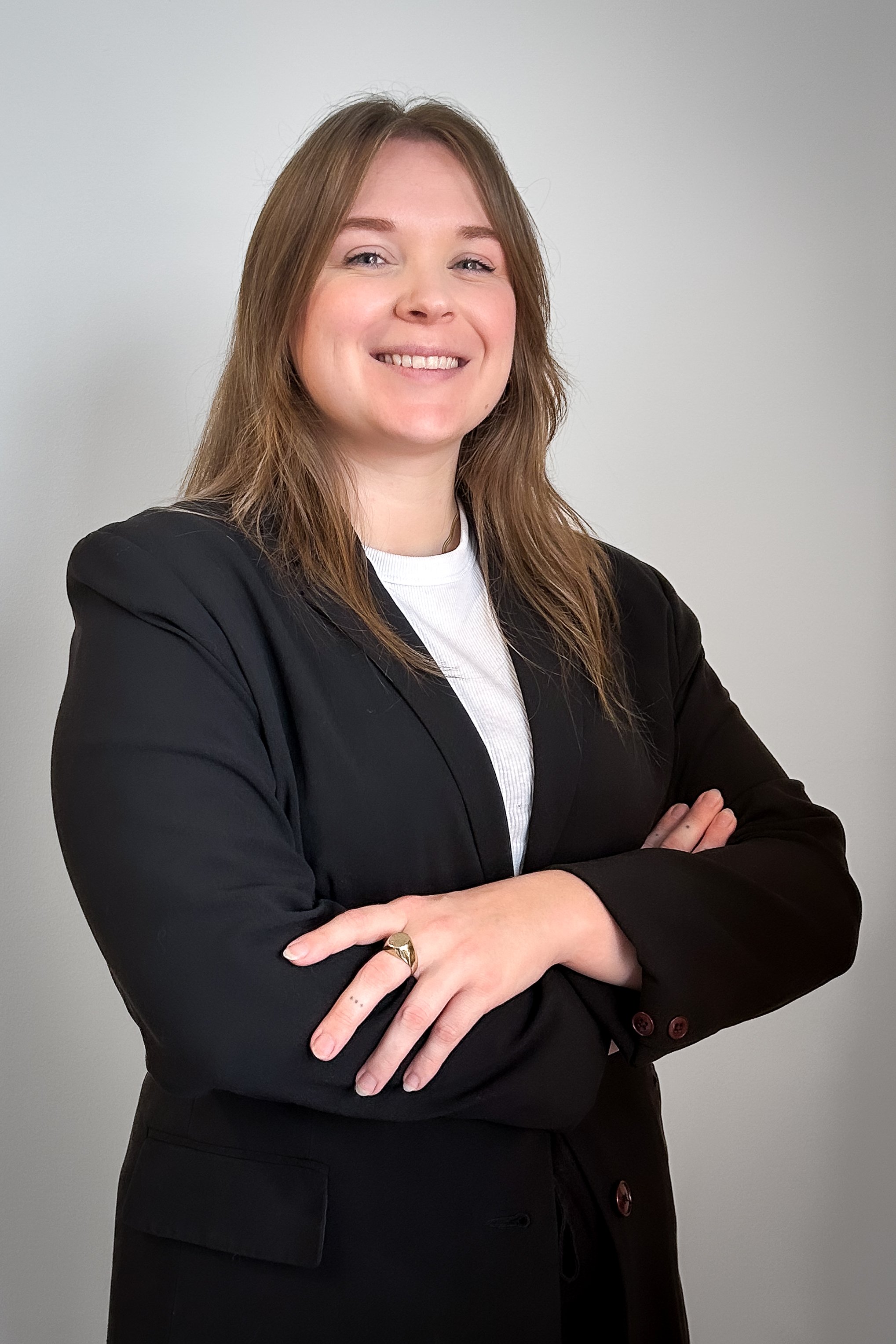 Portrait of Beth Burns smiling confidently with arms crossed, wearing a black blazer over a white top against a light neutral background.