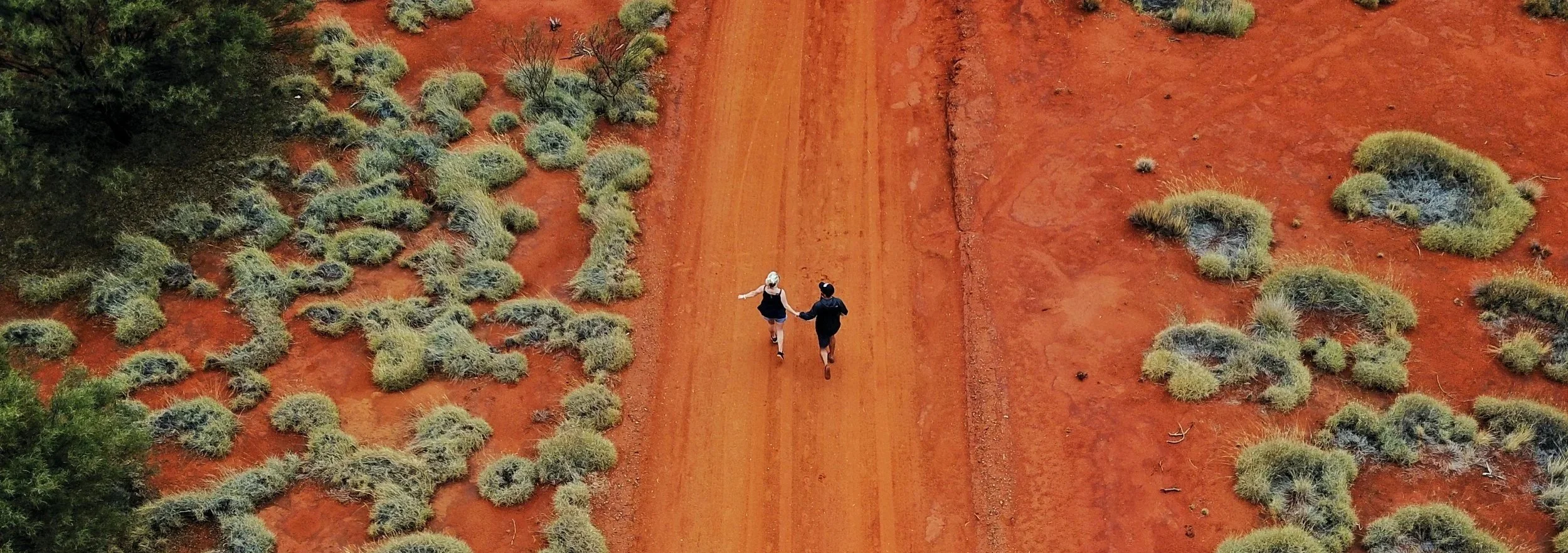 Two people walk hand in hand along a red dirt outback road bordered by scattered scrub, conveying connection, exploration and the vastness of the Australian landscape.