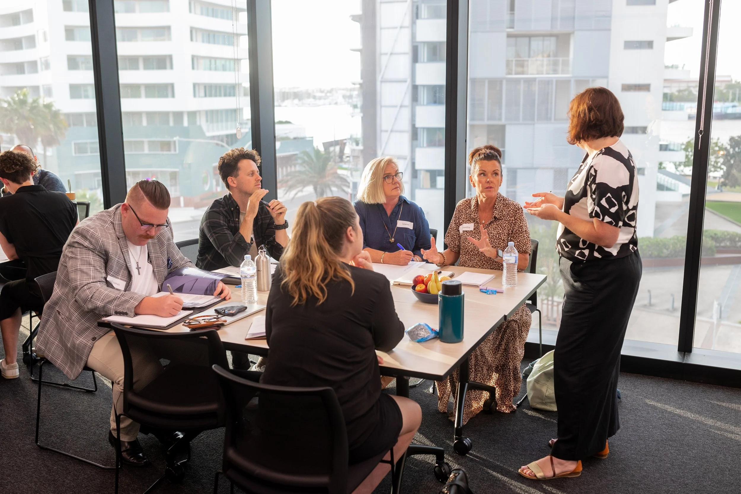 A facilitator guides a small group discussion around a table in a light-filled meeting space, encouraging shared insights, active listening and practical problem-solving in a collaborative workshop setting.