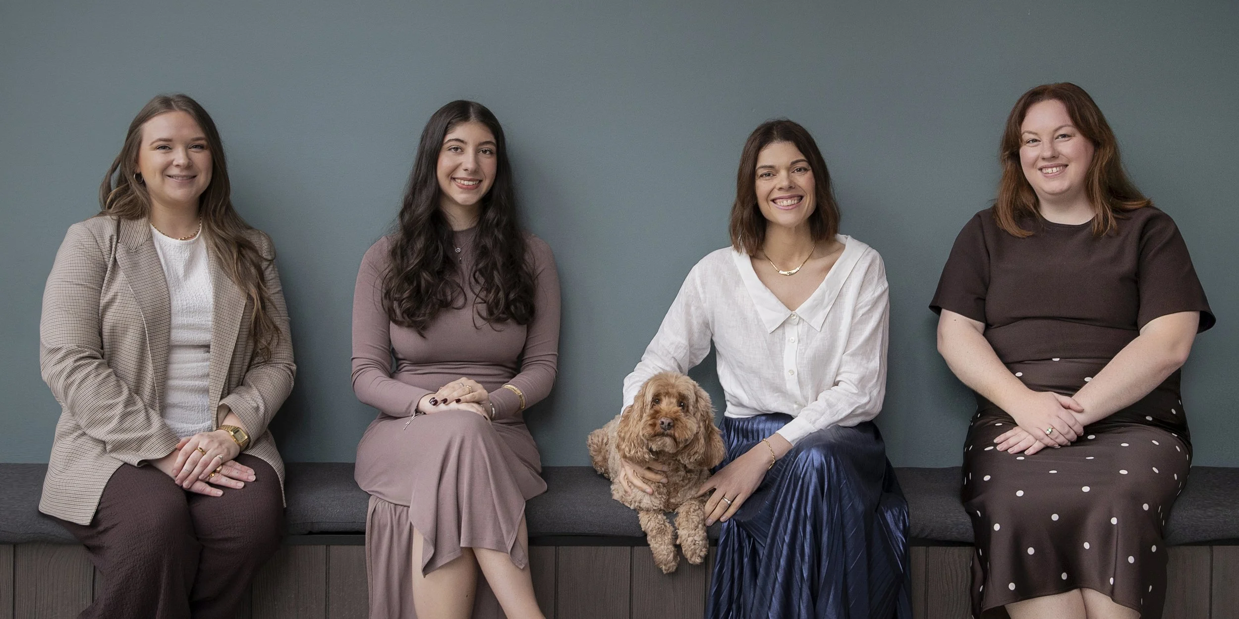 Four smiling women seated together with a small brown dog against a blue-grey wall.