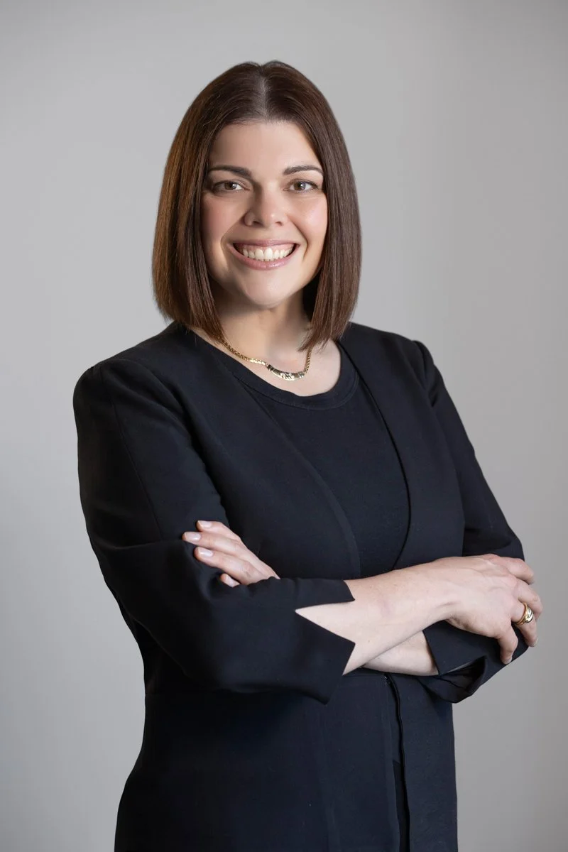 Portrait of Giovanna Lever smiling with arms crossed, wearing a black blazer against a light neutral background.