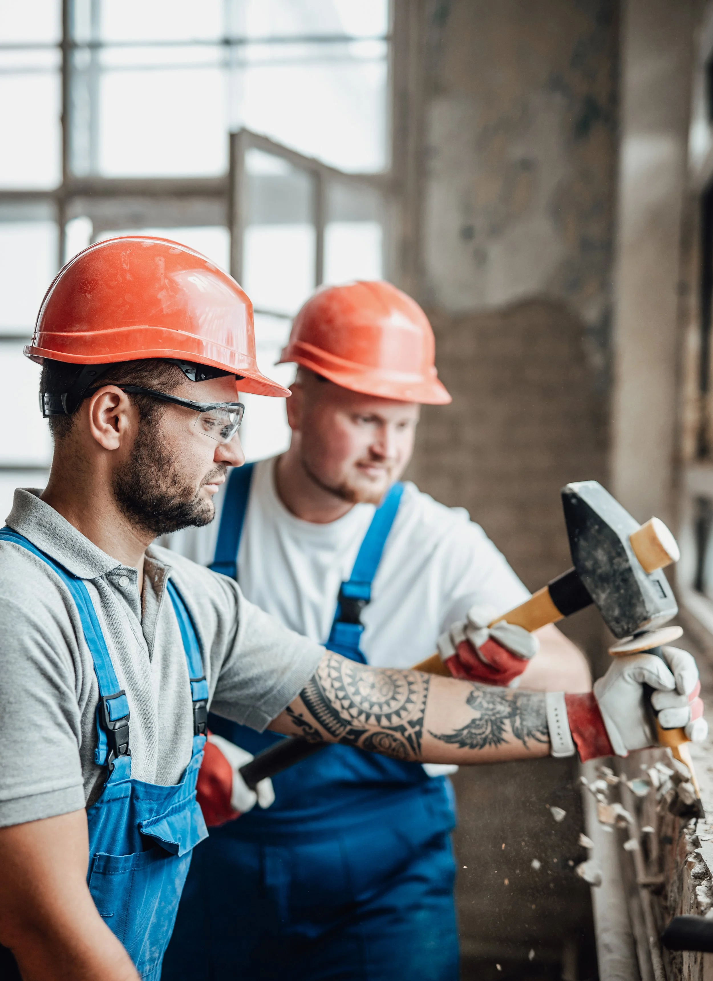 Two builders wearing hard hats work together to remove plaster.