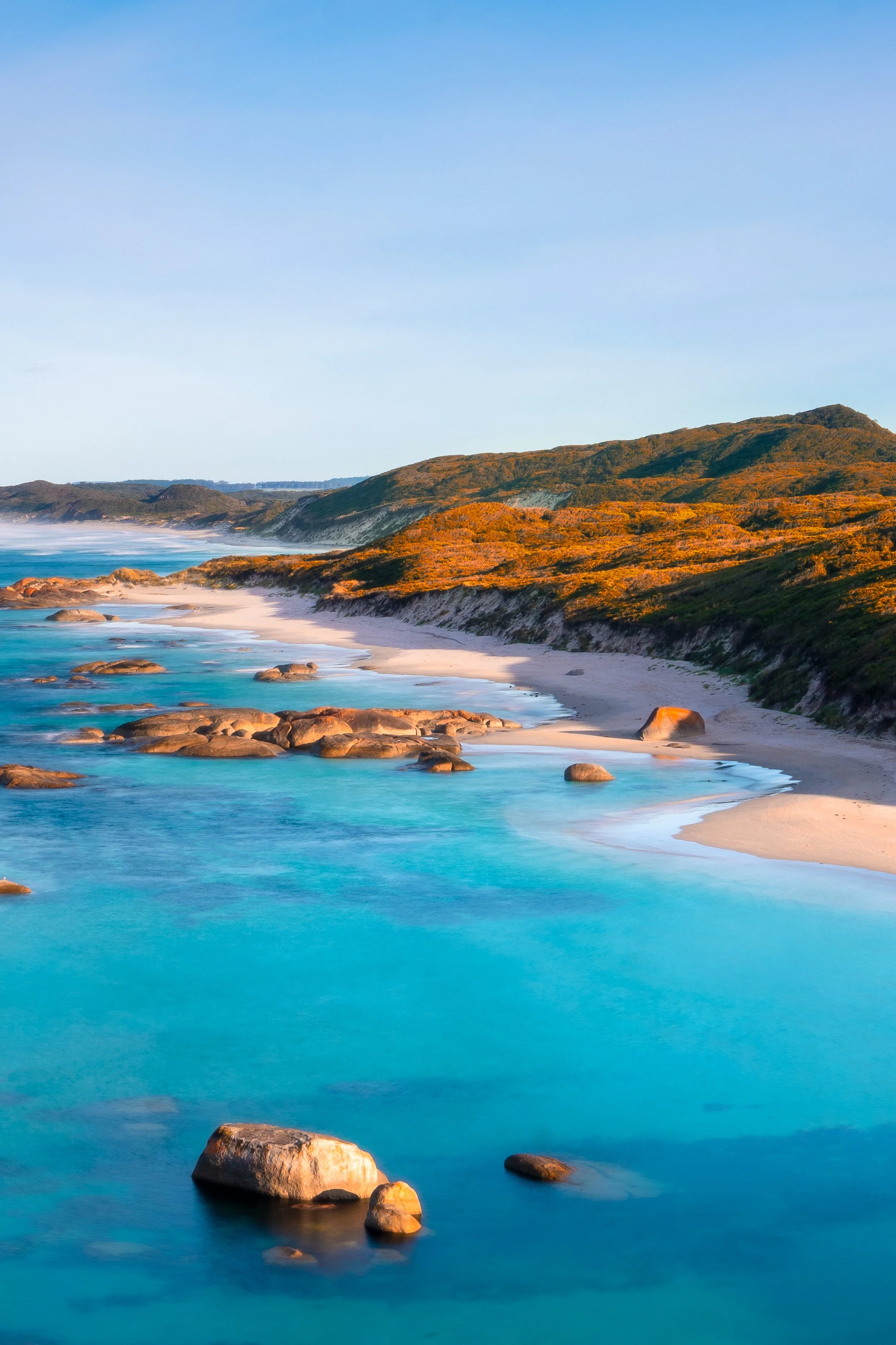 Turquoise water laps against smooth granite boulders and white sand, framed by golden coastal heath and distant headlands beneath a clear blue sky.