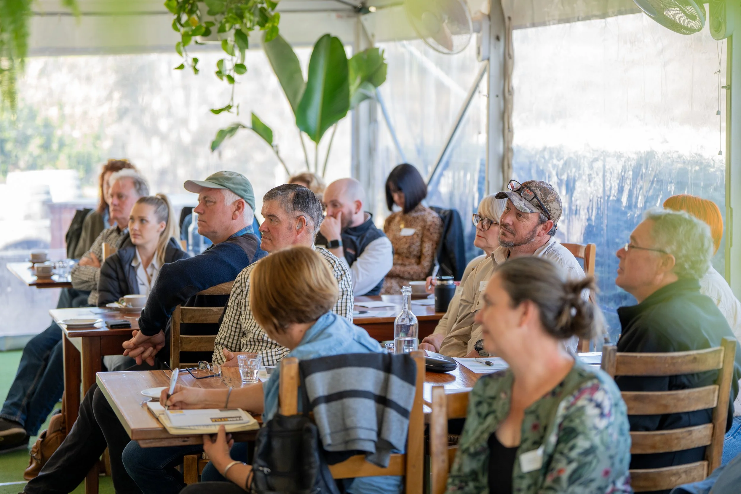 Participants sit attentively in a workshop setting, listening and taking notes.