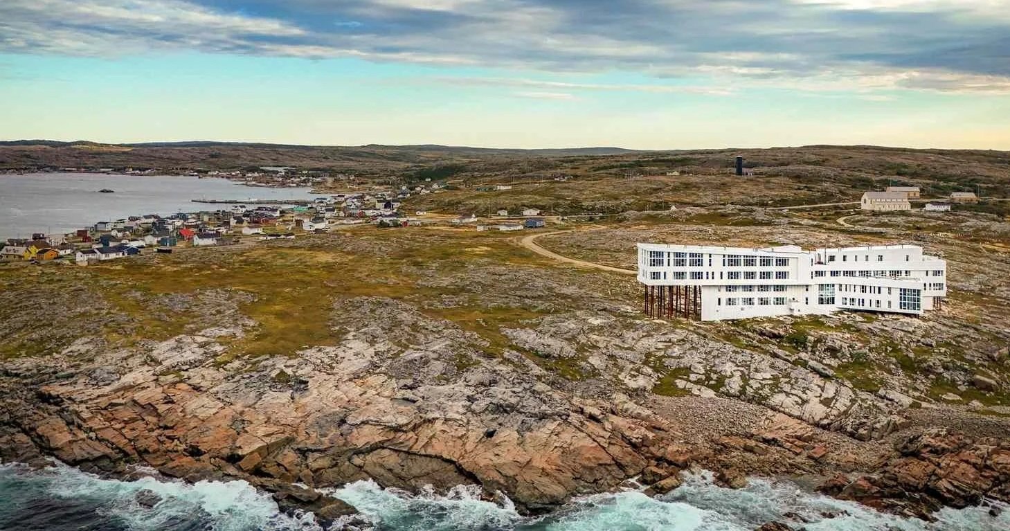 Aerial view of Fogo Island Inn perched on a rocky coastline in Newfoundland, Canada, overlooking the Atlantic Ocean and a small coastal village beneath a cloudy sky.
