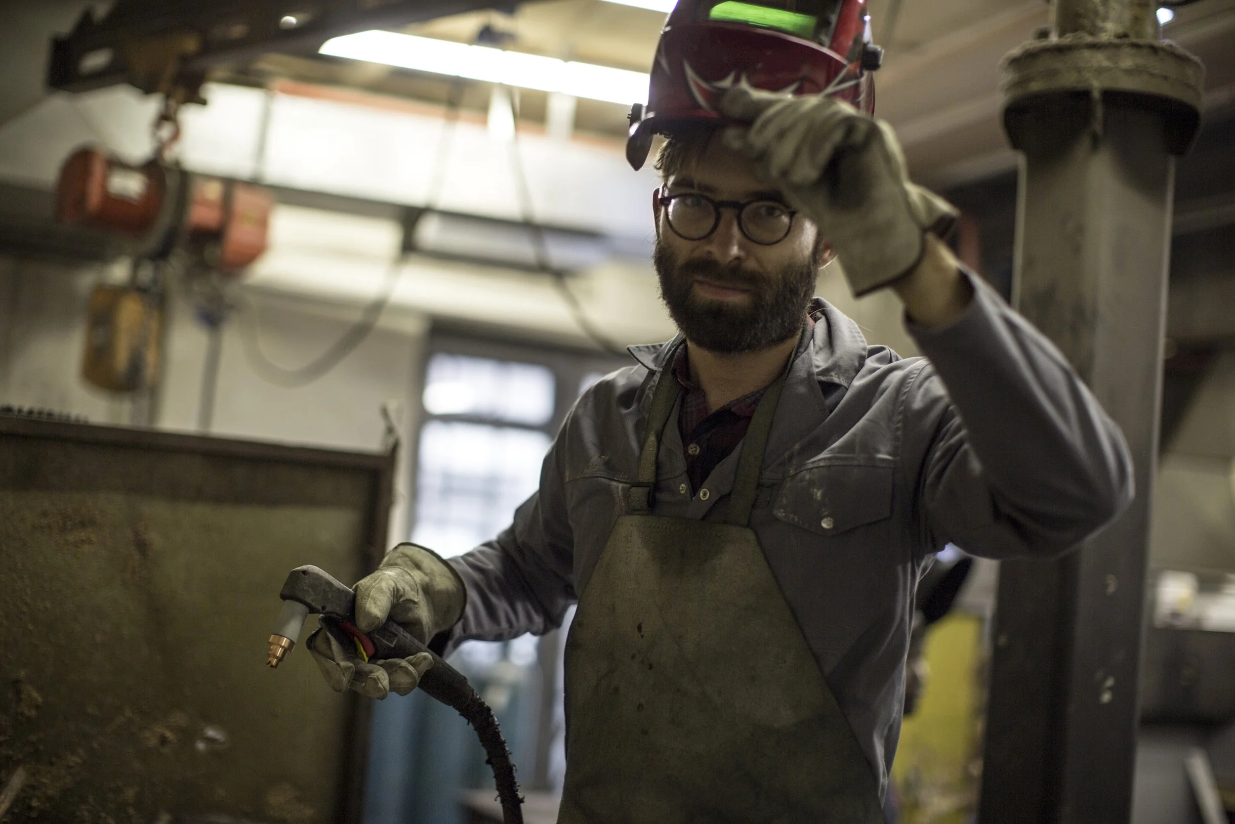 A tradesman in a workshop lifts his welding helmet. He wears protective gloves and a heavy apron in a workshop.