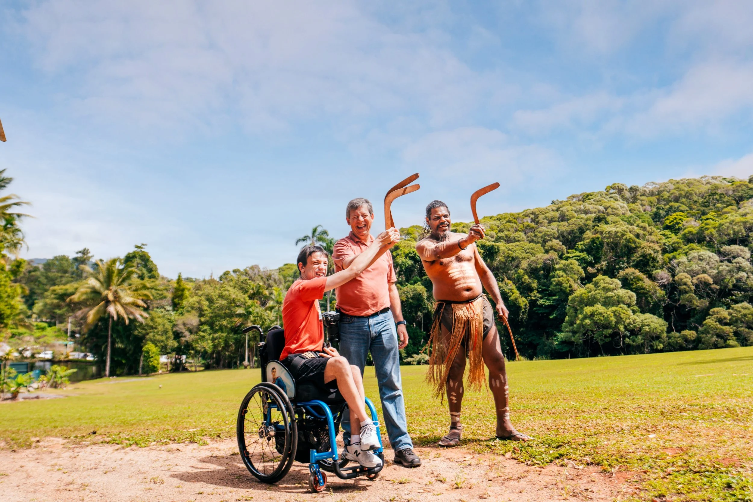 Three men share a joyful outdoor cultural experience as they throw boomerangs together, including a wheelchair user.