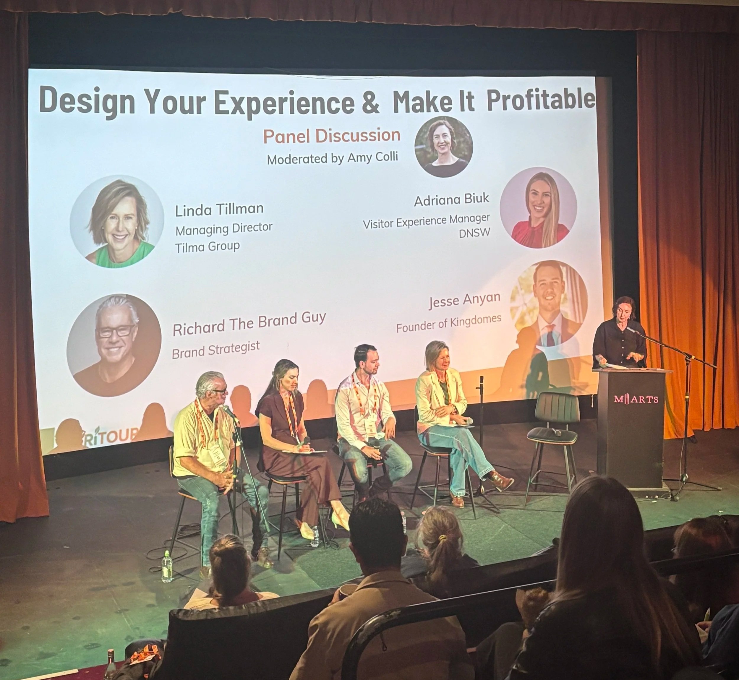 Four panellists seated on stage beside a moderator at a lectern during “Design Your Experience & Make It Profitable”, with an audience in the foreground.