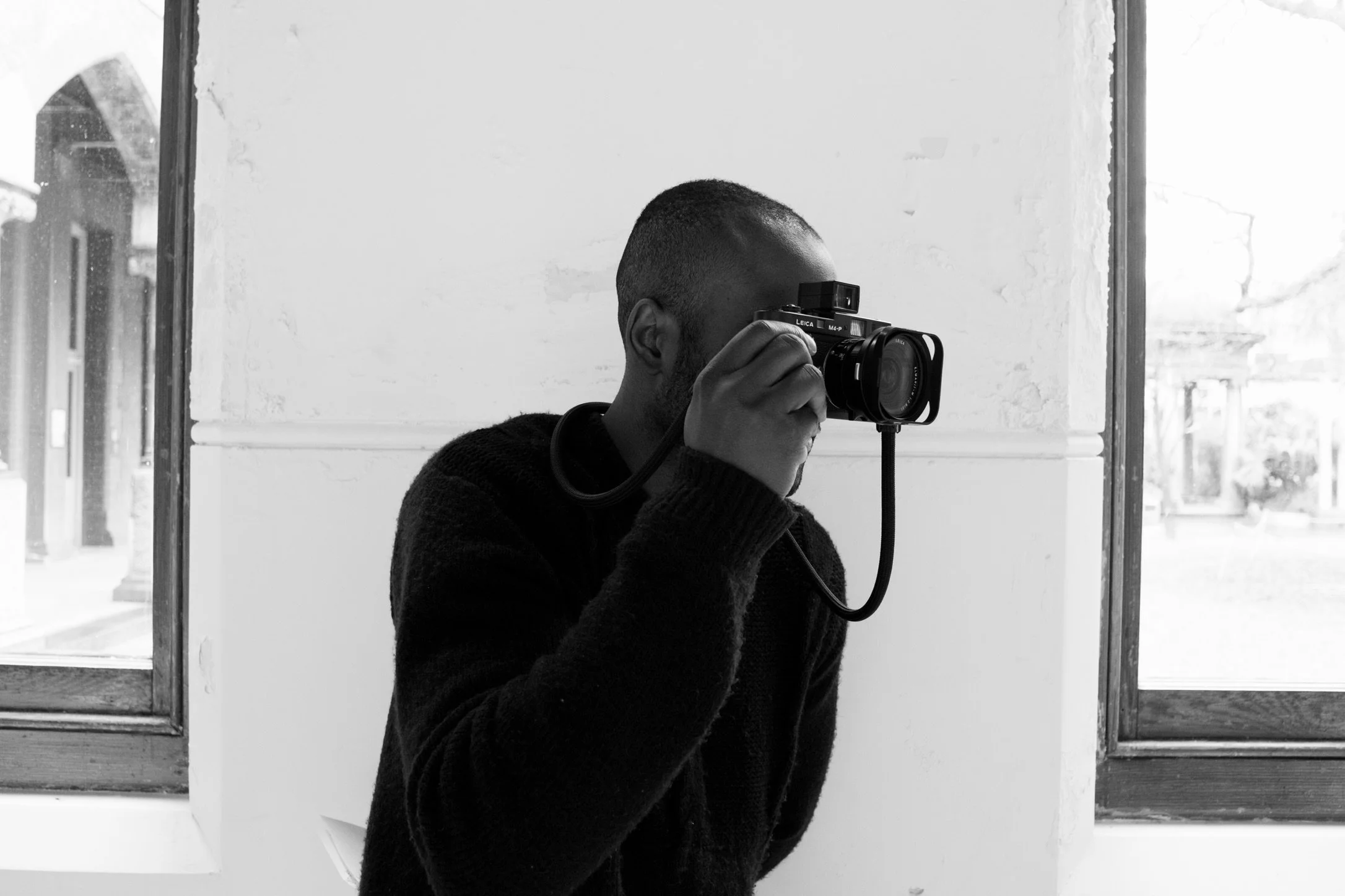 A person, Alain Gratious with a beanie and jacket taking a photograph with a camera while sitting on a jagged rock formation overlooking a foggy valley and mountain range. Leica Camera. Leica M4P
