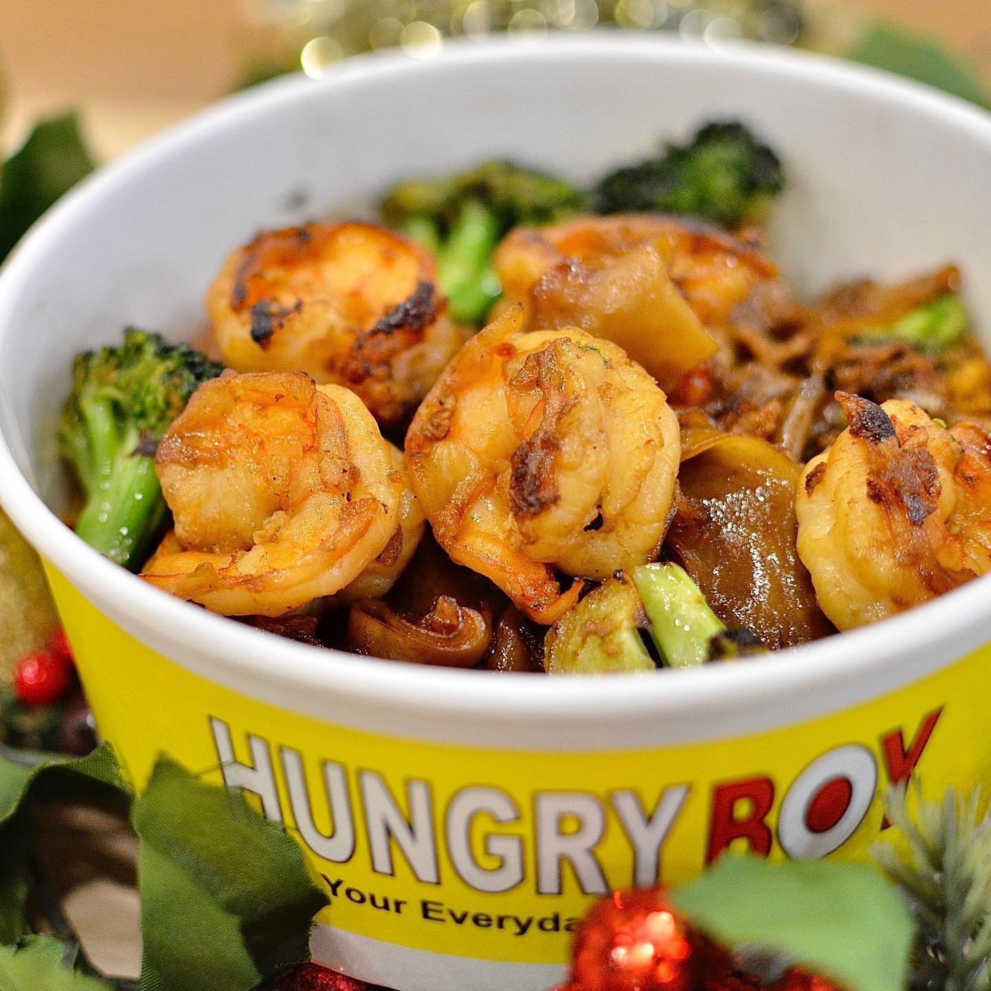 Close-up of a yellow container labeled "Hungry Box" filled with cooked shrimp and mixed vegetables including broccoli, mushrooms, and bell peppers.