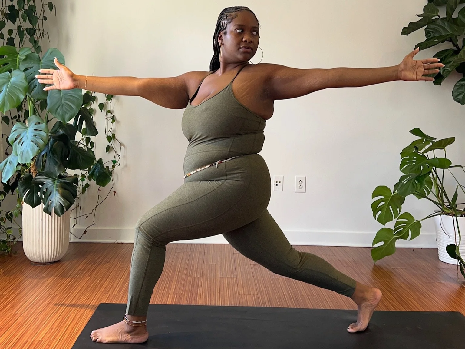 A Black woman practicing yoga indoors, standing in a Warrior II pose with arms extended and looking to her right. She is wearing a matching green athletic outfit, barefoot, on a black yoga mat, with large potted plants on either side.