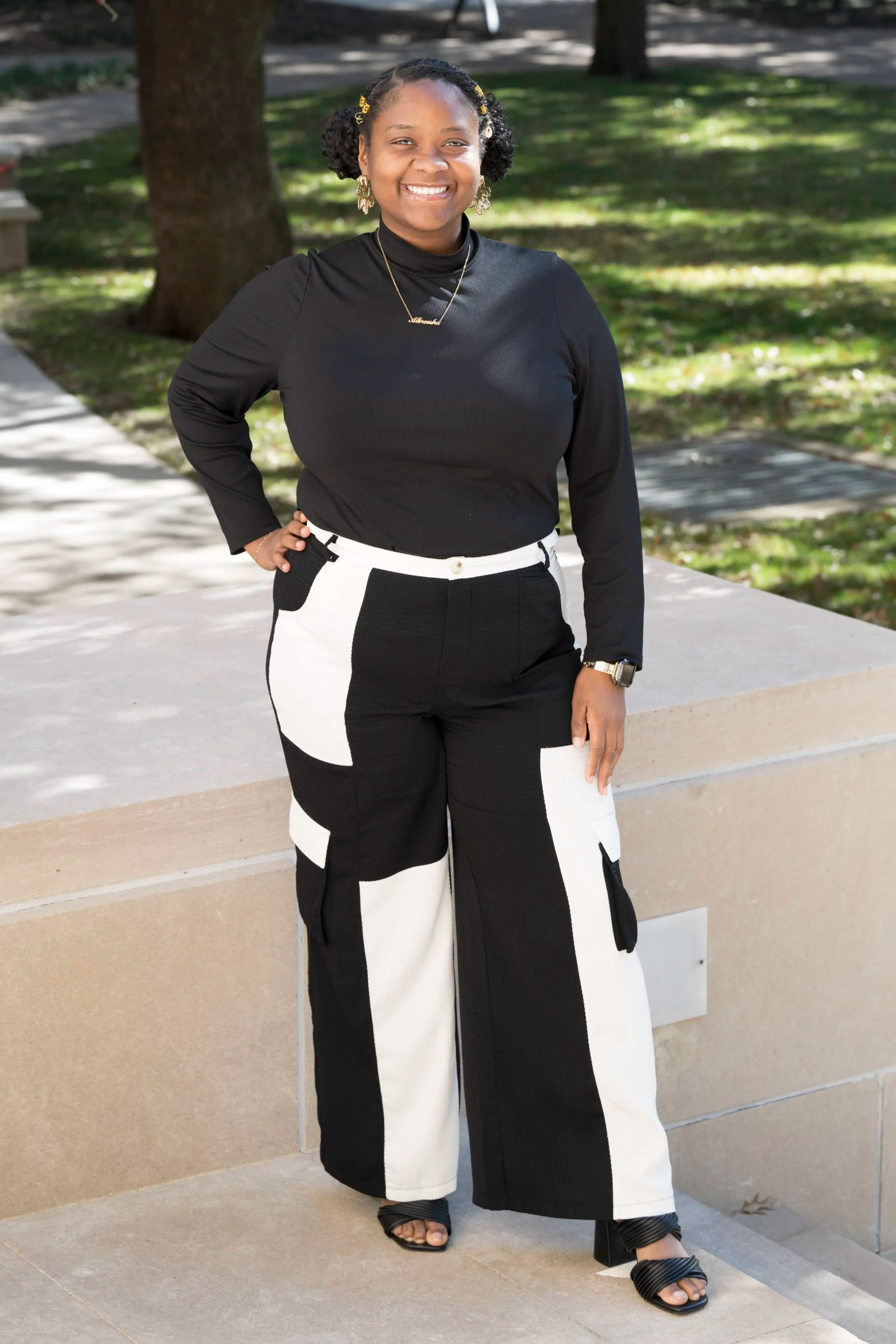 A woman standing outdoors on a concrete ledge in a park, smiling, with trees and grass in the background. She is dressed in a black long-sleeve top, black and white pants with patch pockets, black high-heeled sandals, and wearing jewelry, including earrings, a necklace, and a watch.