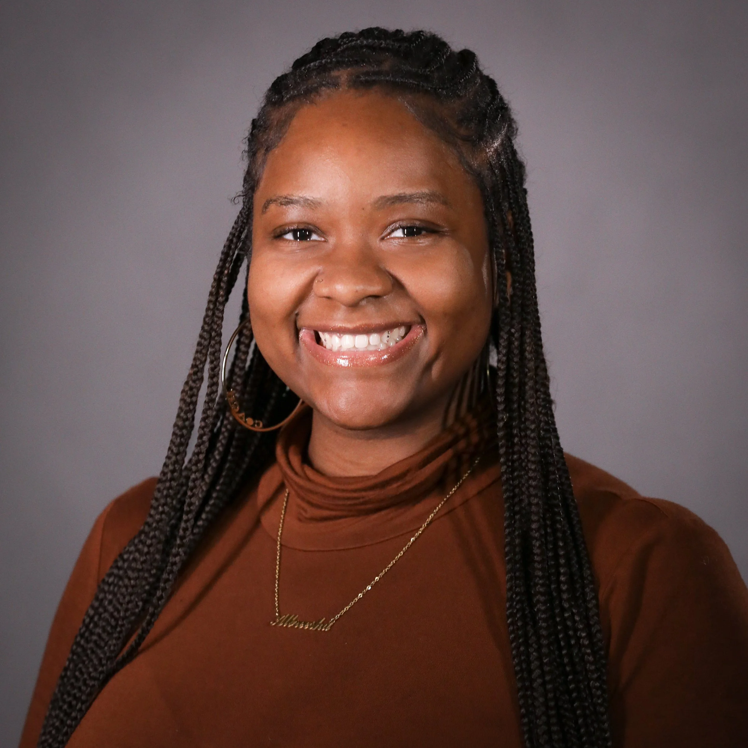 A Black woman with braided hair smiling, wearing a brown top, hoop earrings, a gold necklace, and a friendly expression.