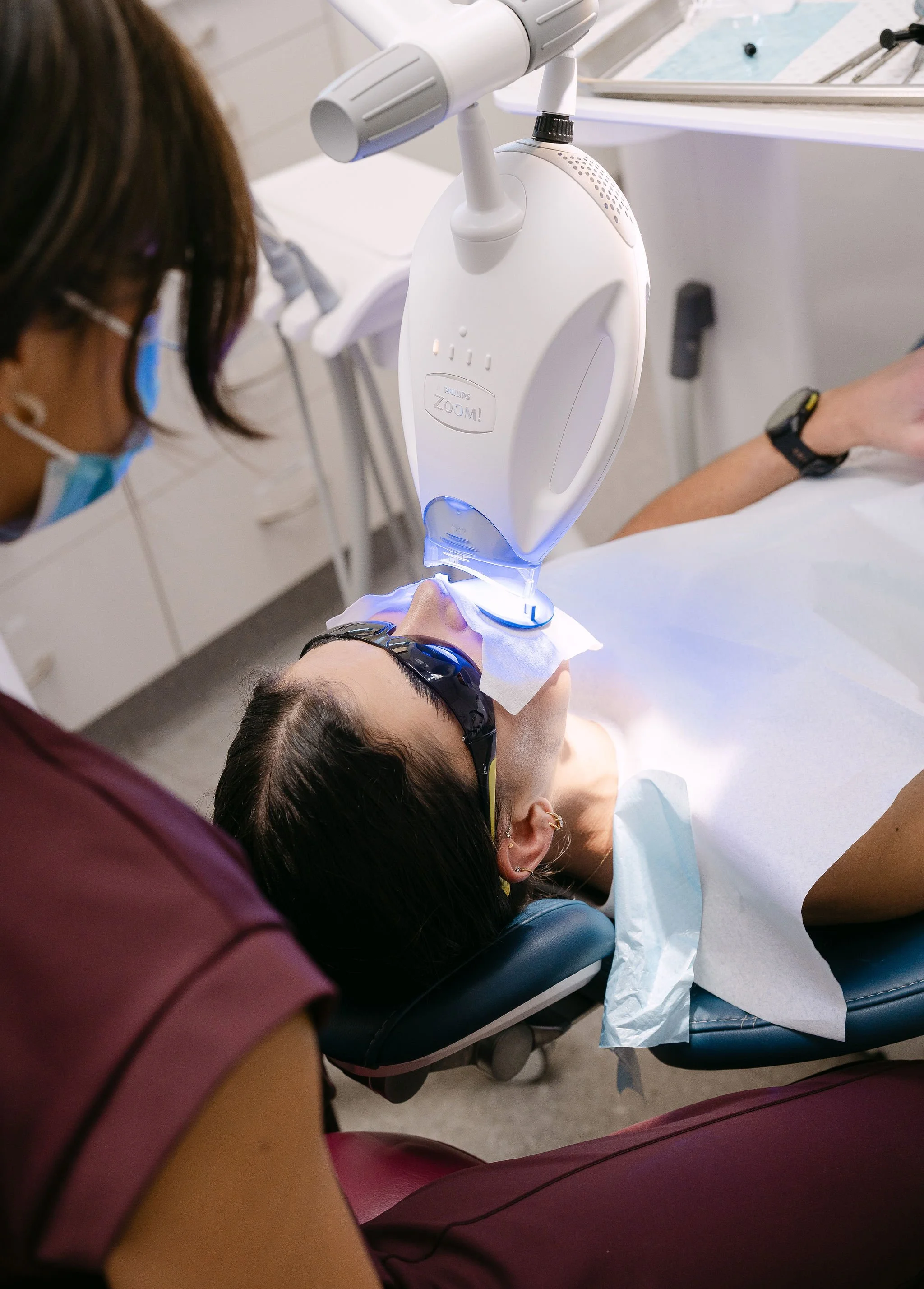 A woman is receiving dental treatment with a curing light, wearing protective goggles and lying on a dental chair in a clinic.