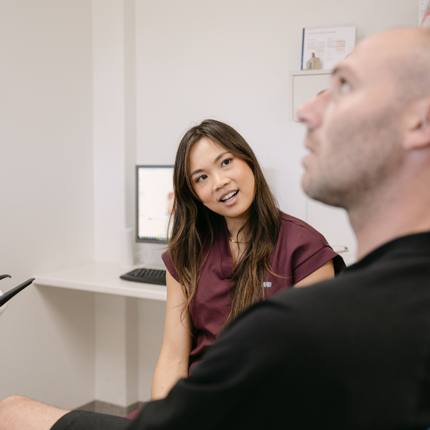 A woman in a maroon medical uniform talking to a man in a black shirt in a clinical office setting.