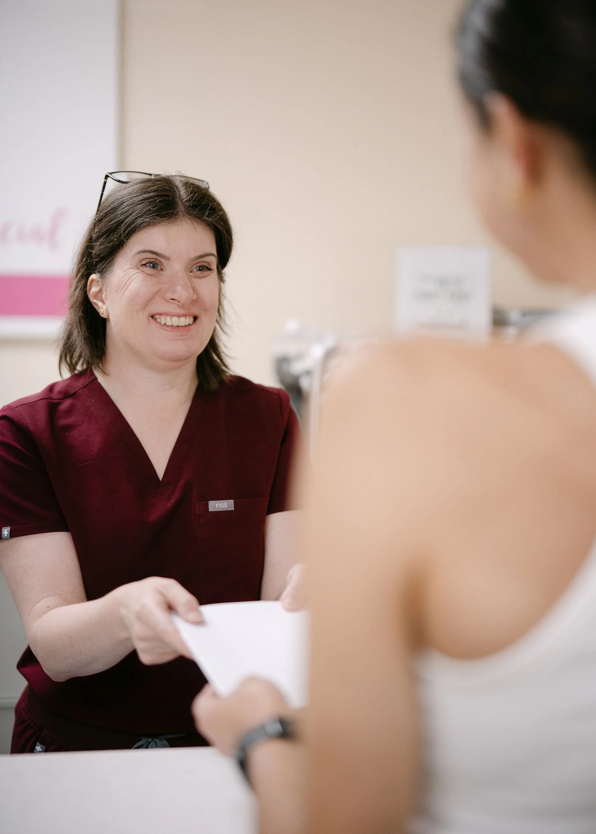 A nurse in medical scrubs smiling as she hands papers to a patient in a healthcare setting.