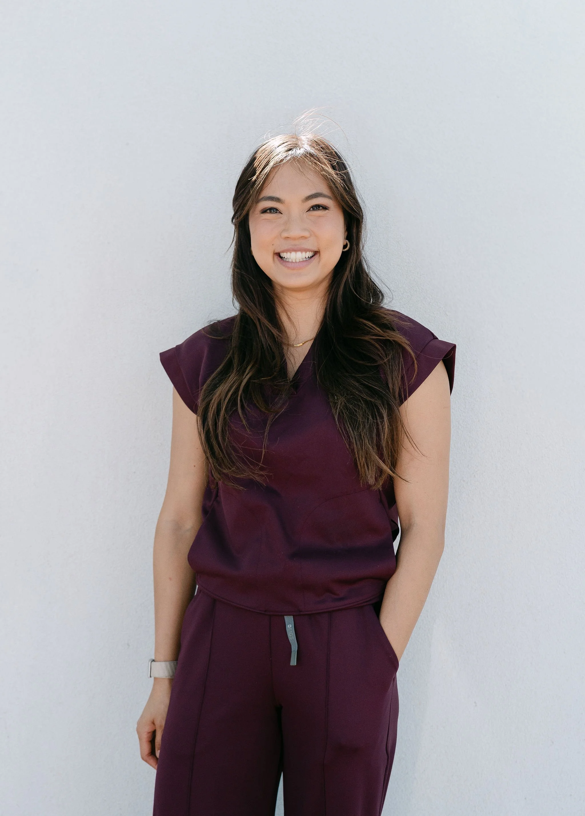 Smiling woman with long dark hair wearing a purple outfit standing against a white wall.