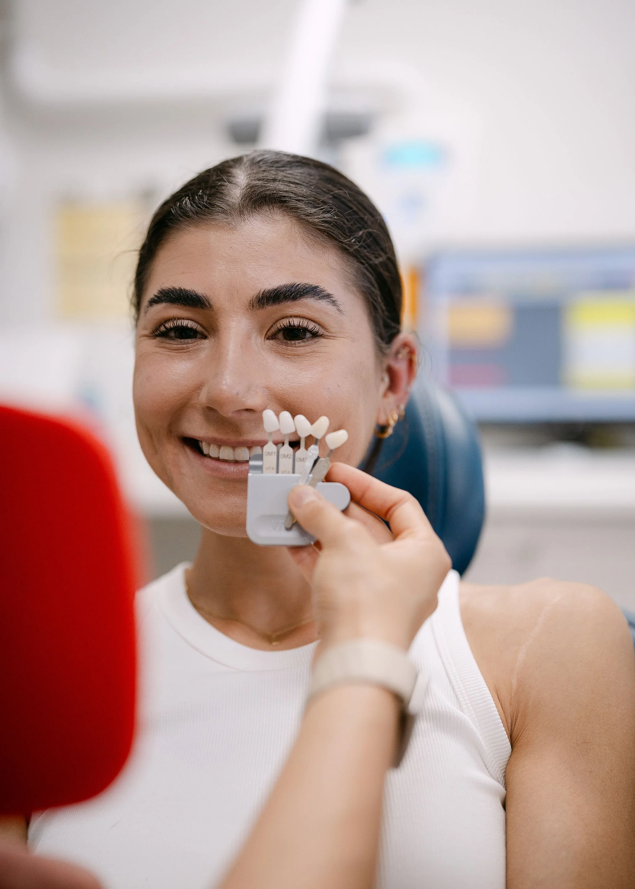 A woman smiling at the dentist during a teeth whitening procedure, holding a shade guide with various tooth color samples.