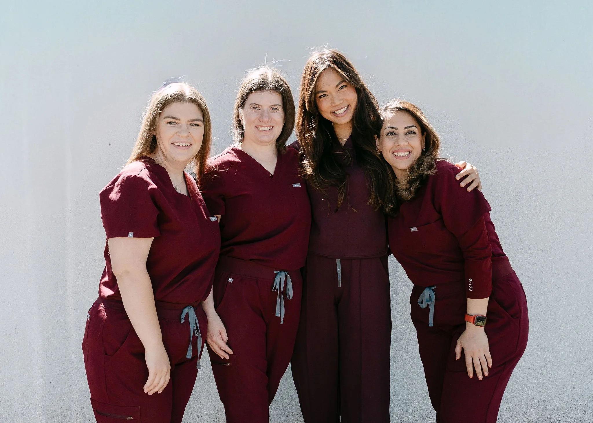 Four women in matching maroon scrubs smiling and standing together outdoors against a light gray wall.