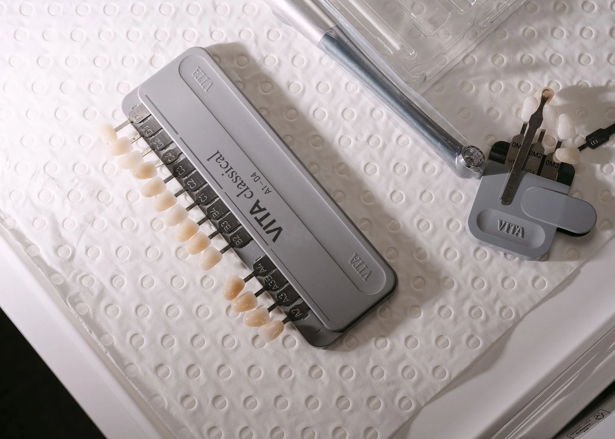 A dental shade guide with various tooth color samples on a metal strip placed on a white textured surface, with a dental instrument stand nearby holding dental tools.