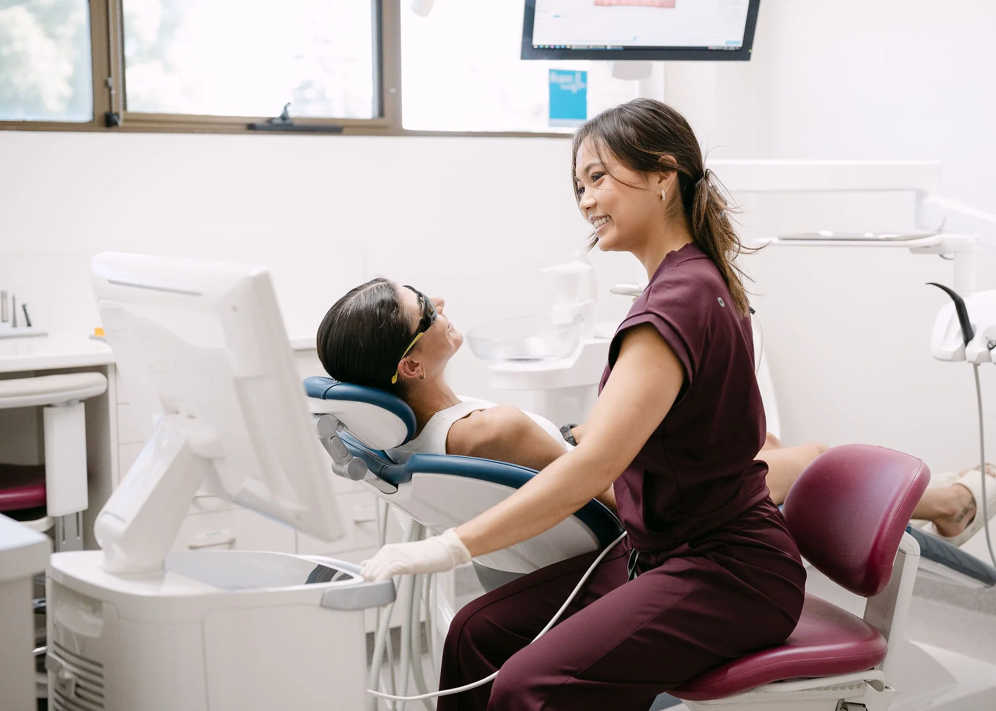 Dental hygienist smiling at a patient lying in a dental chair, inside a bright dental clinic.