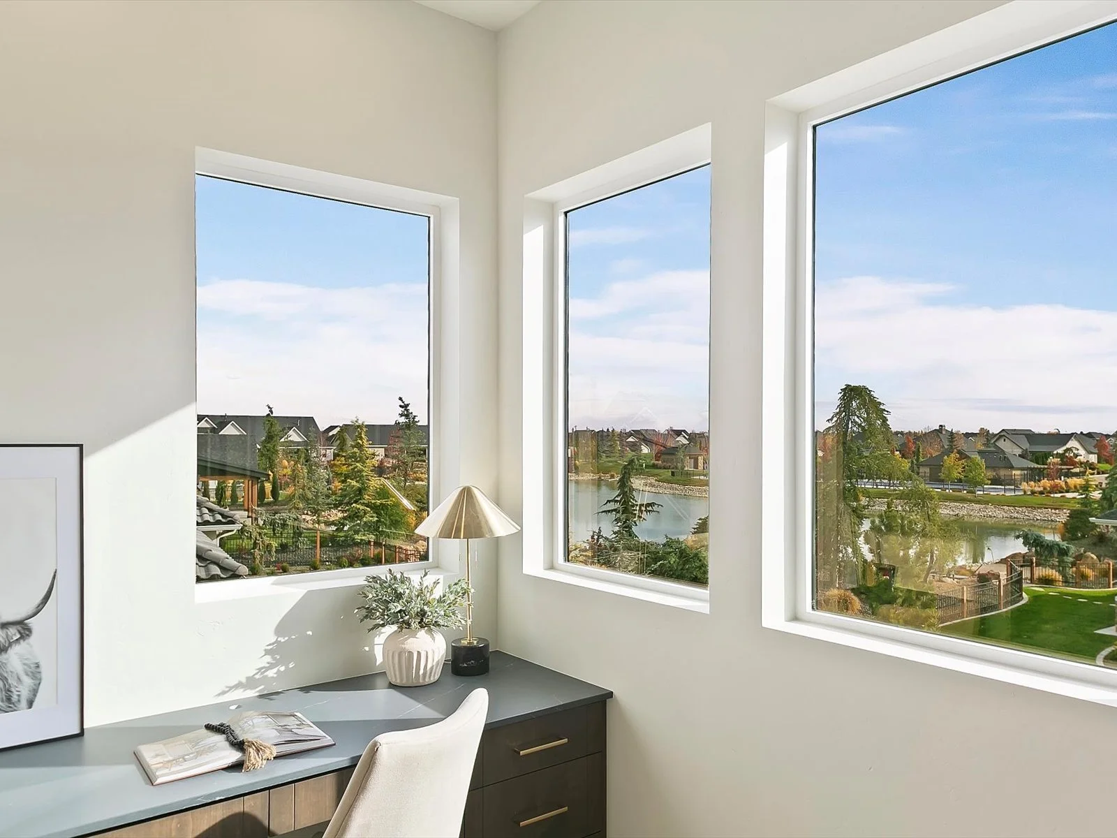 A corner of a bright home office with three large windows overlooking a neighborhood with houses, trees, and a pond, decorated with a desk, a chair, a framed photo, a potted plant, a lamp, and an open book.