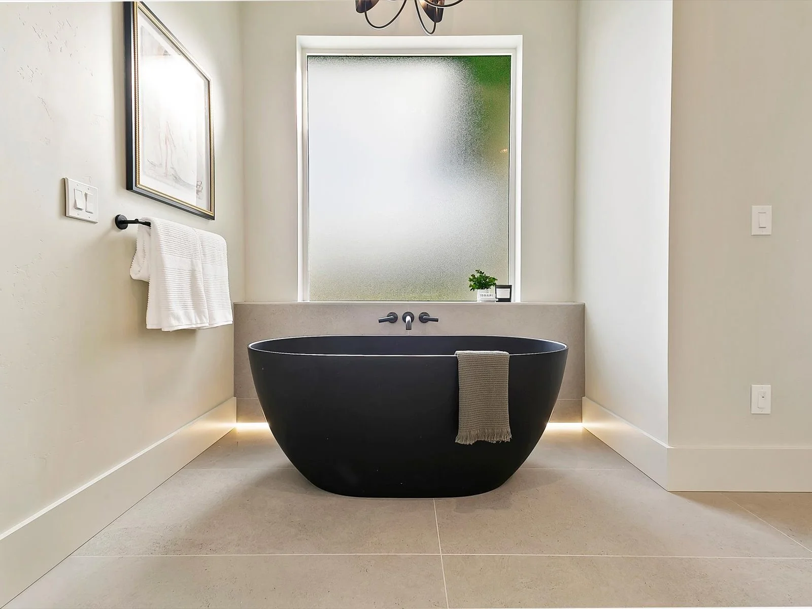 Modern bathroom with a black freestanding bathtub, frosted window behind, small plant, towel hanging on the tub, and artwork on the wall.