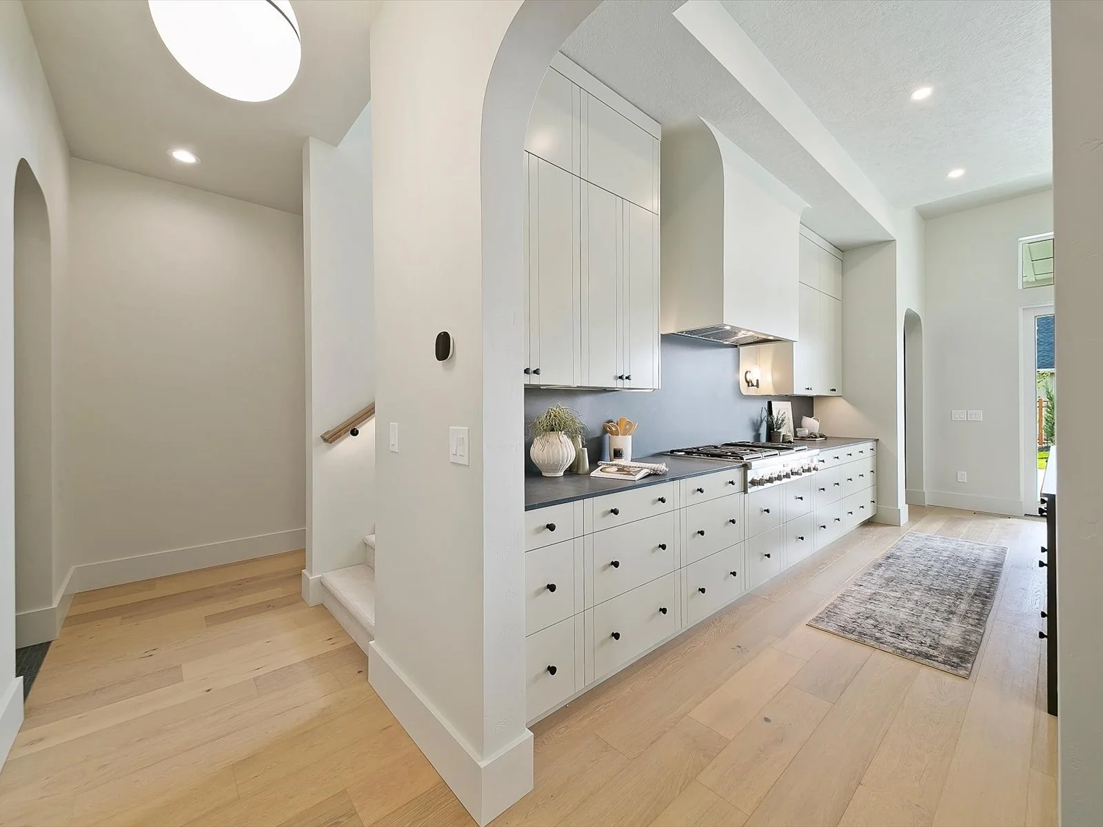 Modern kitchen with white cabinets, black knobs, a gray backsplash, and a light wood floor. There is a stove, a potted plant, and kitchen utensils on the countertop.