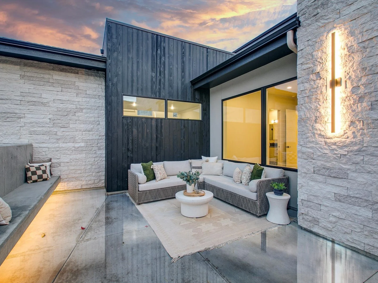 Modern outdoor patio with white sectional sofa, decorative pillows, potted plants, concrete flooring, and wall lighting at sunset.