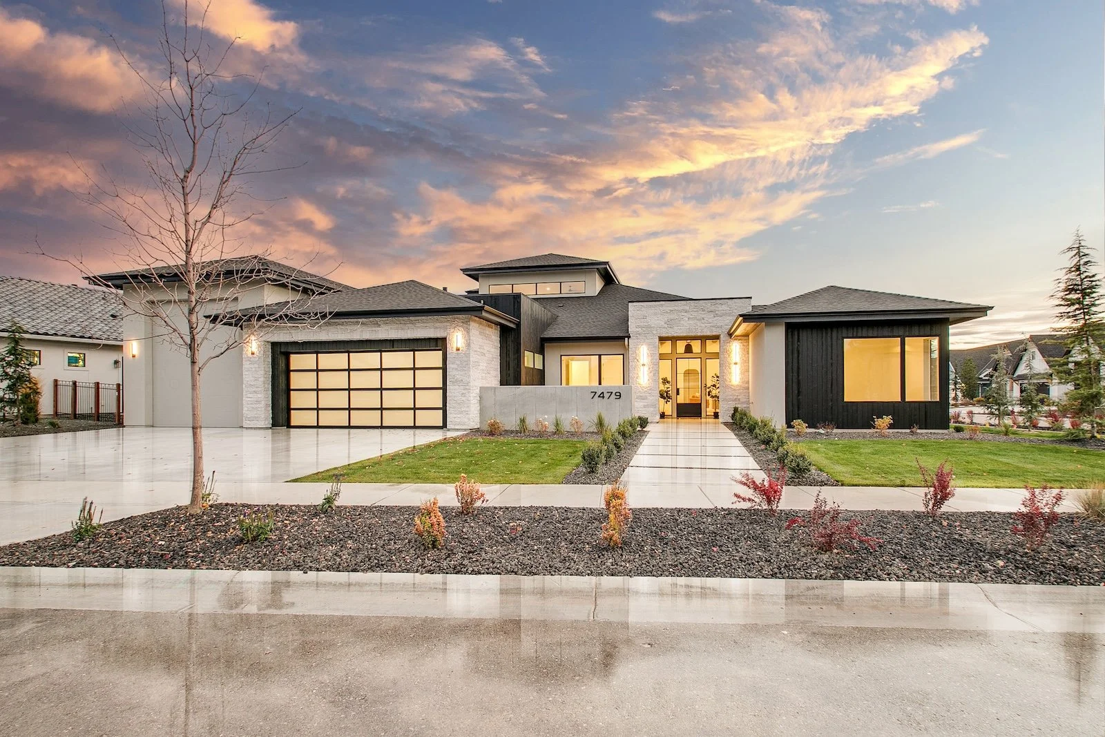 Modern house with a brick and black exterior, large windows, and a two-car garage at sunset, with a manicured lawn and walkway leading to the front door.