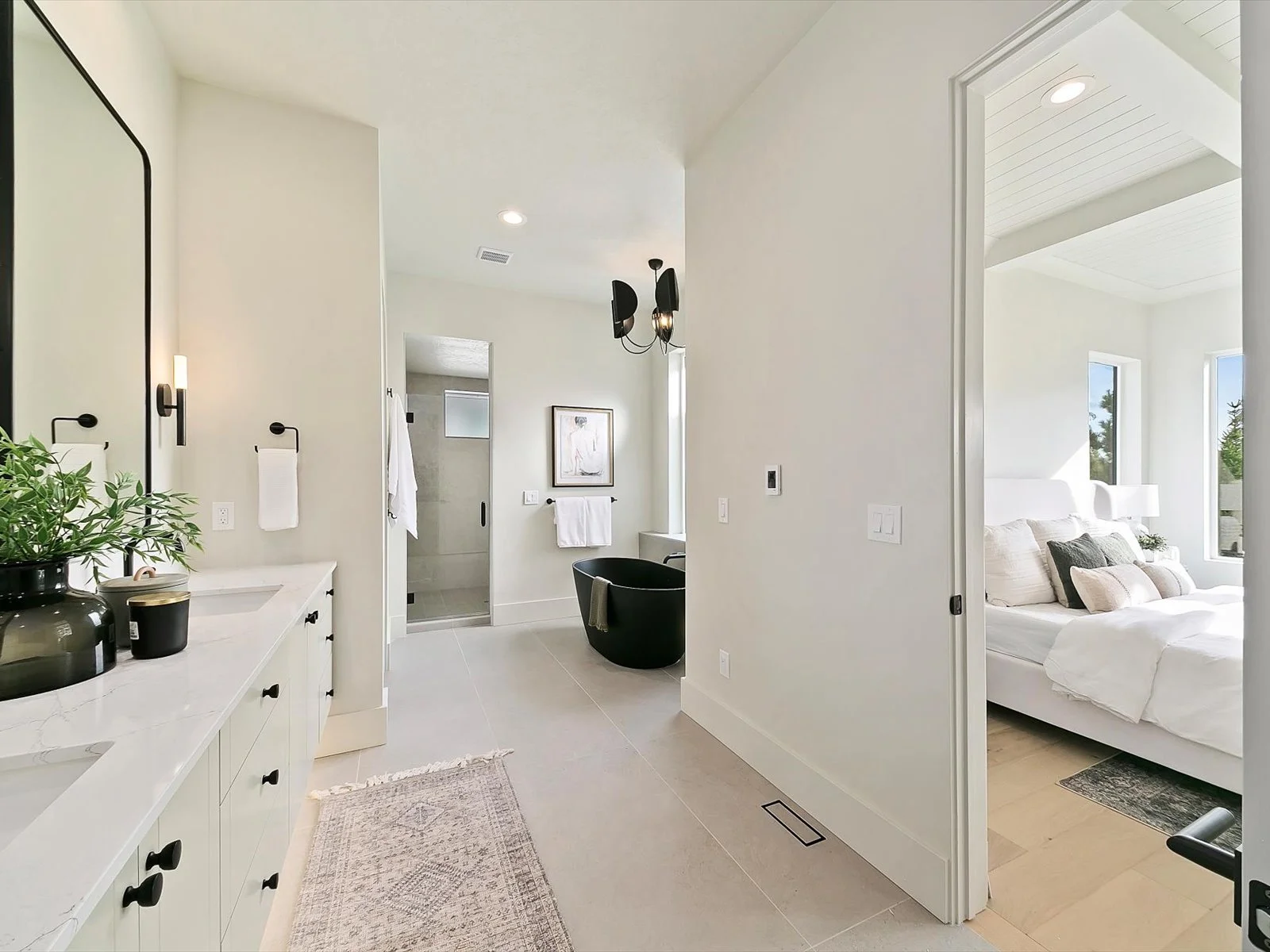 Modern bathroom with a walk-in shower, black freestanding bathtub, white walls, and a bedroom with large windows and white bedding visible through an open door.