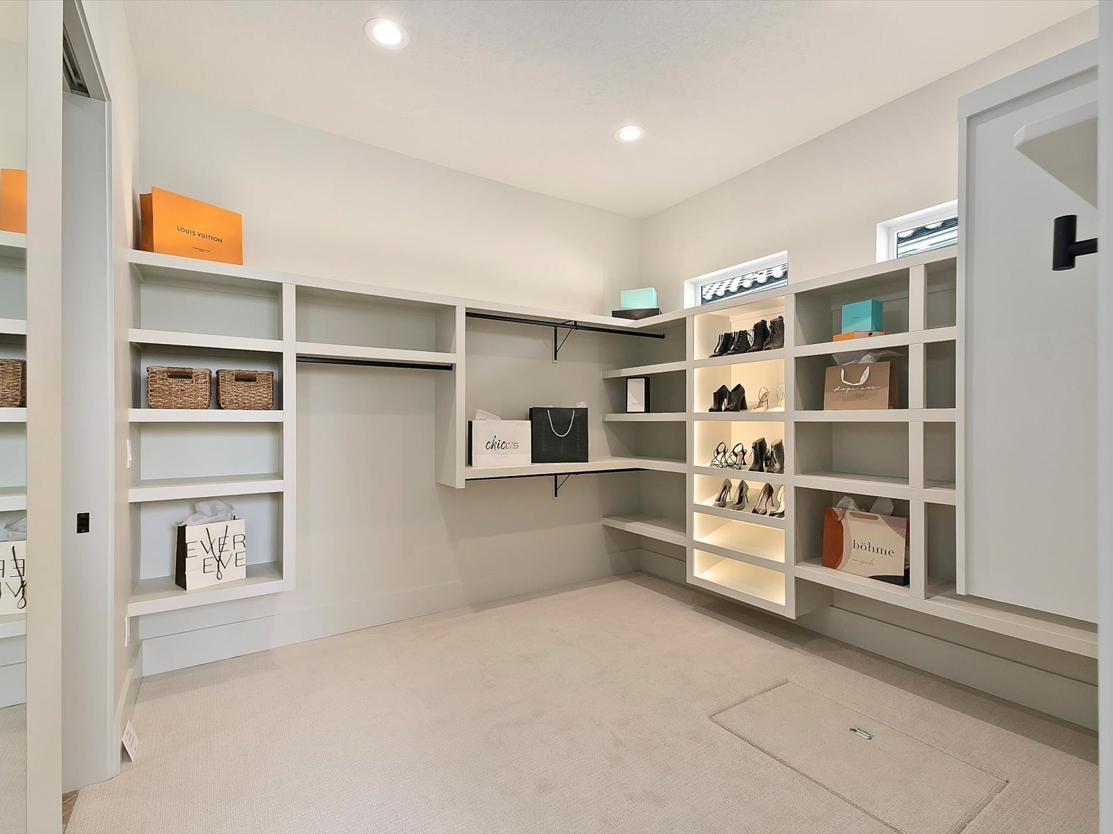 Walk-in closet with white built-in shelves holding shoes, bags, and boxes in a bright room with recessed lighting and small windows.