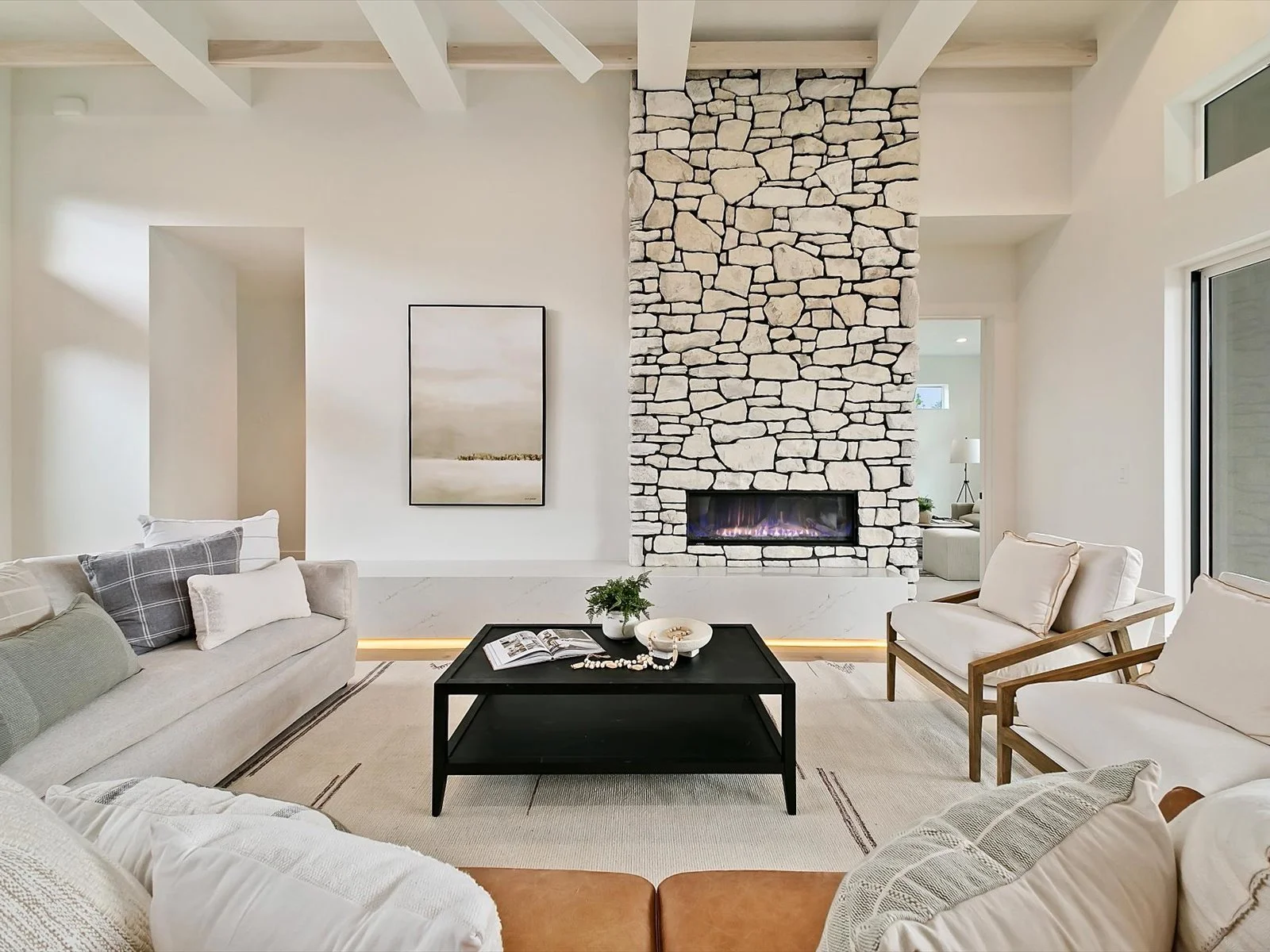 Living room with a large stone fireplace, white walls, beige and white furniture, black coffee table, and artwork on the wall.
