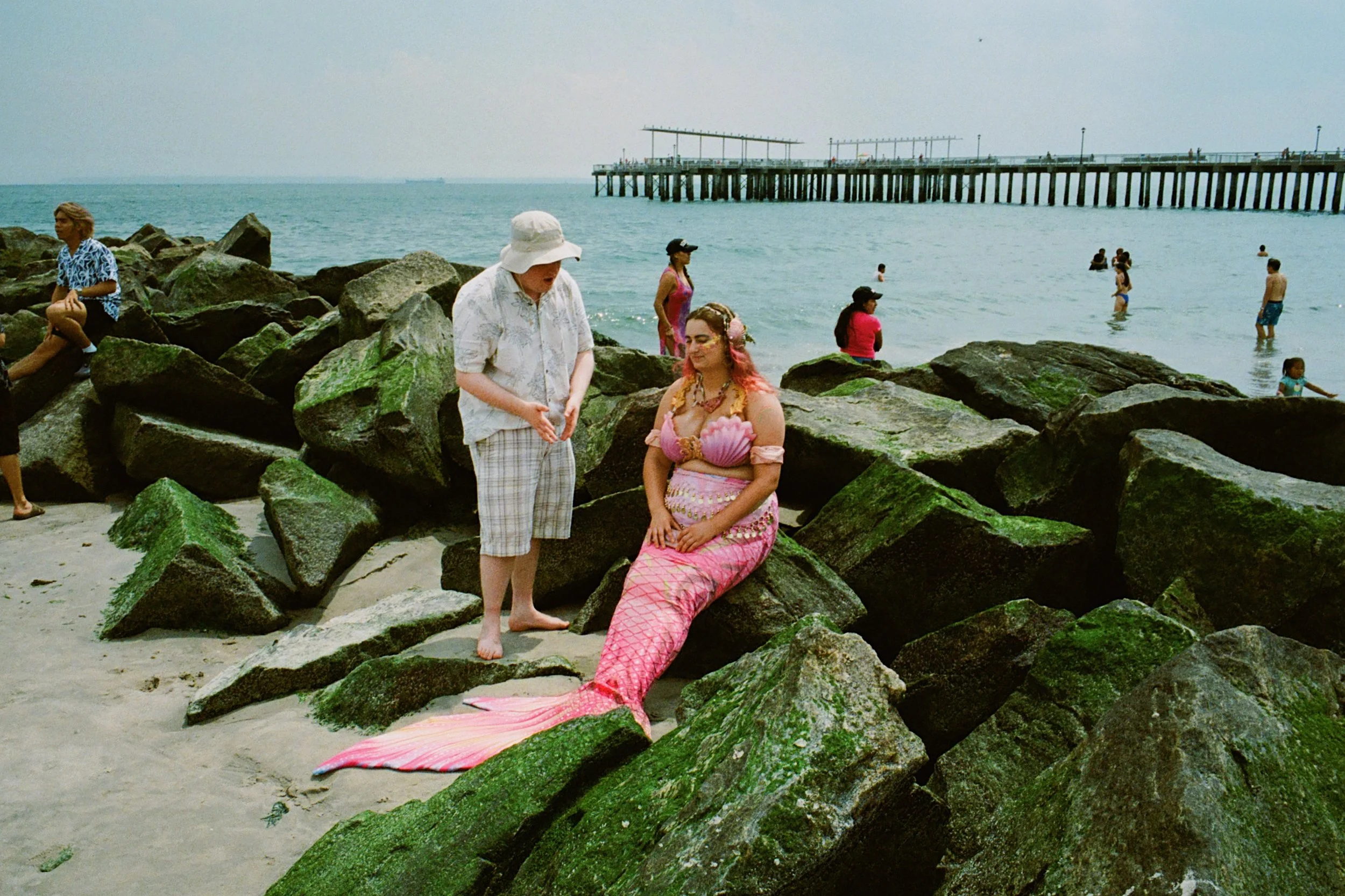 A mermaid dressed in pink sitting on rocks at the beach while a woman stands nearby. People are swimming and walking along the shoreline in the background, with a pier extending into the ocean.