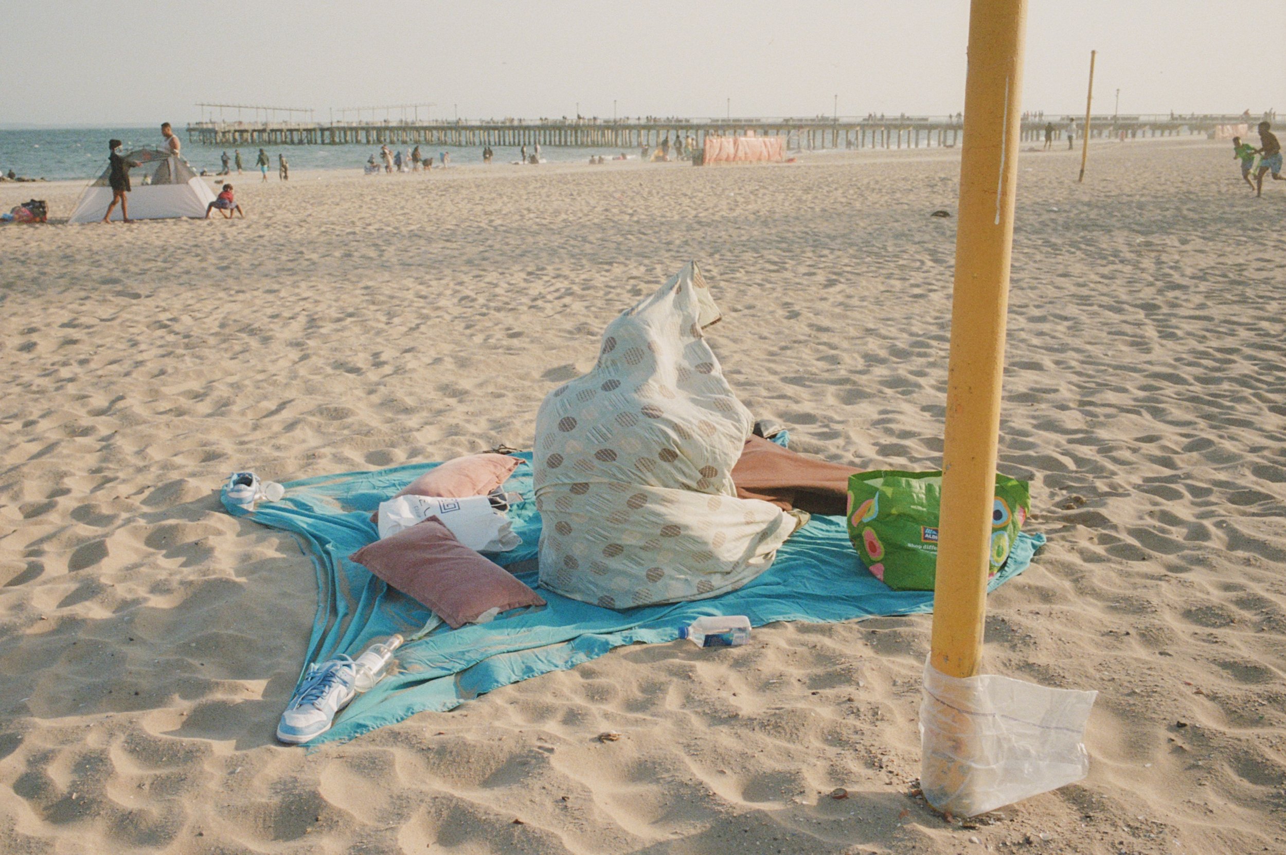 Empty blanket and pillows on the sandy beach near a yellow pole, with a hoodie or blanket draped over a cushion, and a green bag nearby, in a busy beach setting with people and a pier in the background.