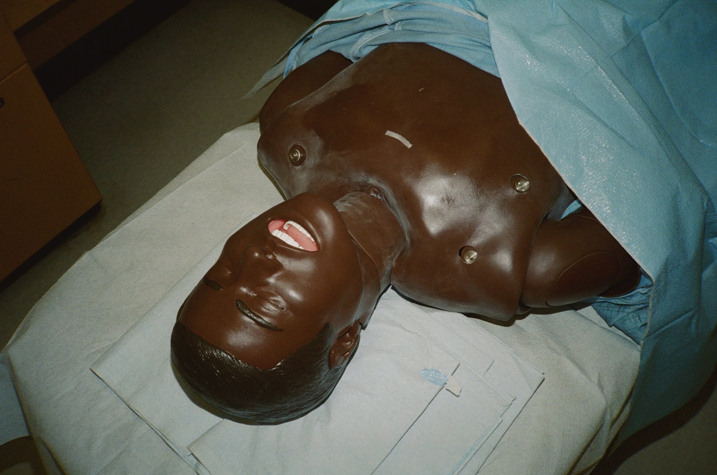 A medical mannequin or model lying on a hospital bed, with a detailed face and chest, simulating a human patient for educational or training purposes.