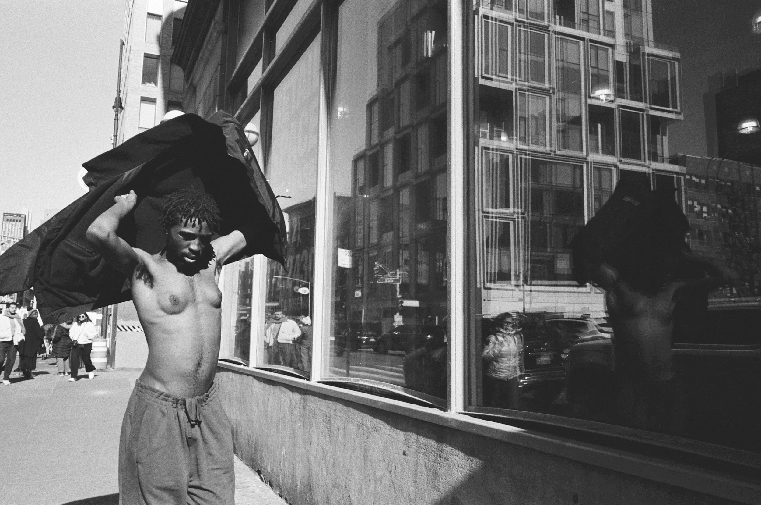 A shirtless man with dreadlocks is putting on a jacket beside a large glass building, with city street and pedestrians reflected in the window.
