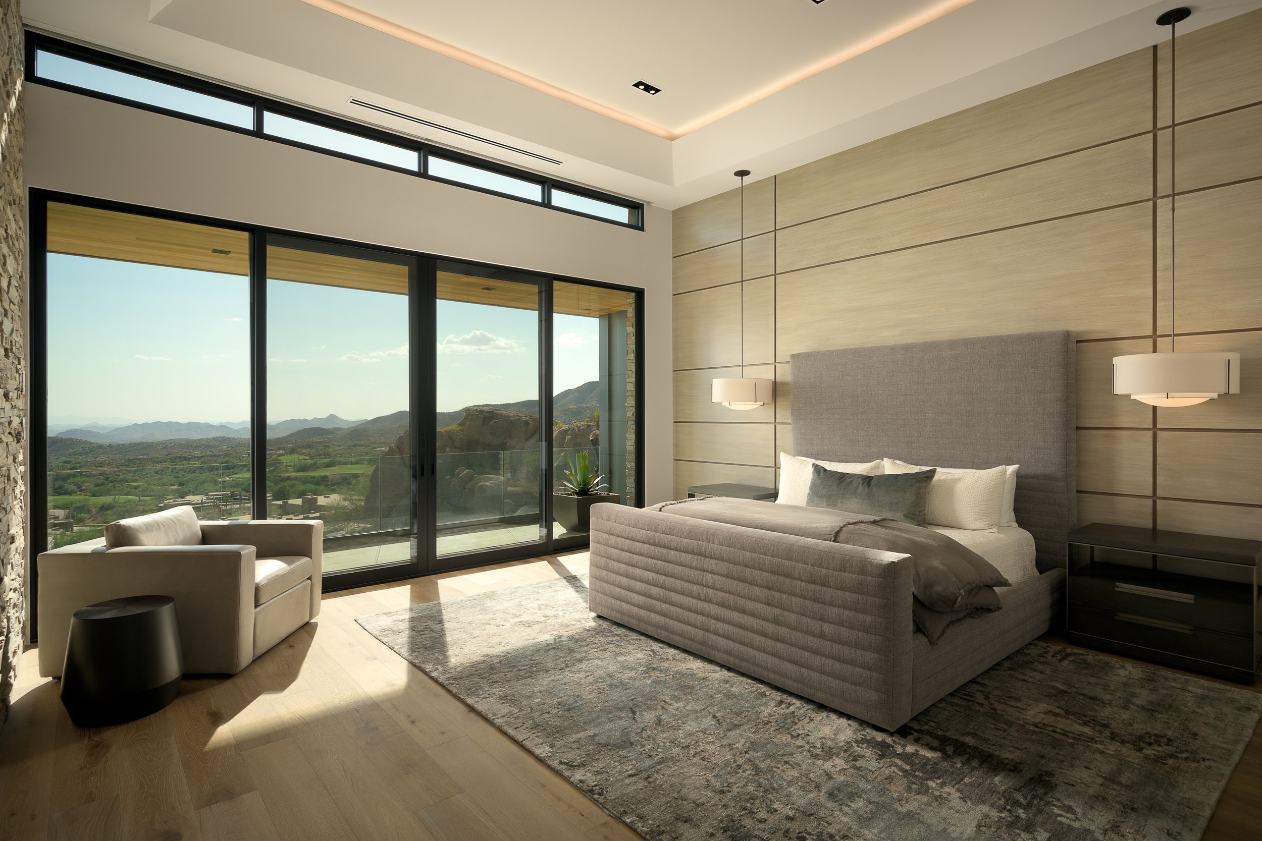 Modern living room with large glass sliding doors opening to a scenic mountain view, a gray upholstered bed with pillows, a leather armchair, a black side table, and hanging pendant lights, with wood flooring and a textured stone wall on the side.